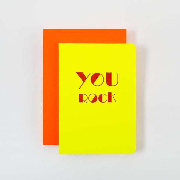 „YOU ROCK Karte und Kuvert“ von Le Typographe besteht aus zwei farbenfrohen Klappkarten auf weißem Hintergrund: Die obere Karte ist leuchtend gelb mit „YOU ROCK“ in fetten roten Buchstaben und darunter befindet sich eine leuchtend orange Karte.