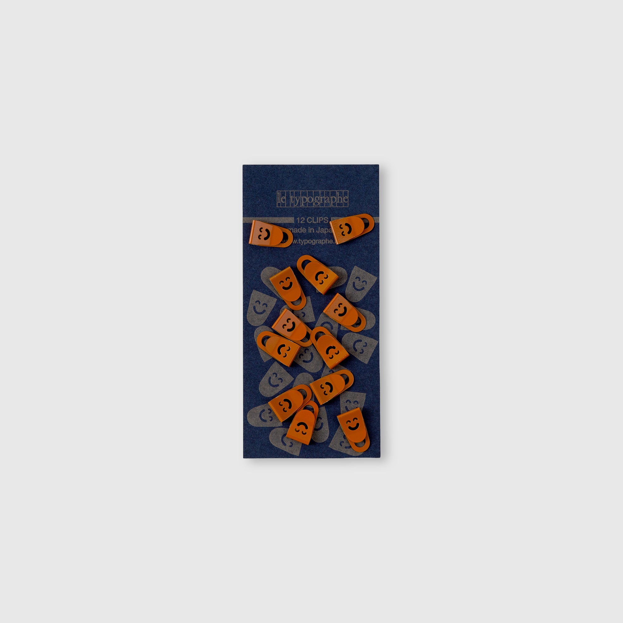 Das Set Mini Clips Smiley von Le Typographe ist auf einer dunkelblauen, rechteckigen Karte angeordnet, die sich perfekt für die Organisation auf dem Schreibtisch eignet. Die Clips in Form eines orangefarbenen und grauen Kürbisses sind mit Smiley-Gesichtern auf hellgrauem Hintergrund versehen.