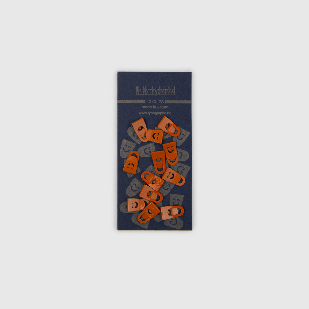 Zwölf orangefarbene Metall-Mini-Clips Le Typographe mit Smiley-Ausschnitten, angeordnet auf einer dunkelblauen, rechteckigen Unterlegkarte zur Organisation auf dem Schreibtisch.