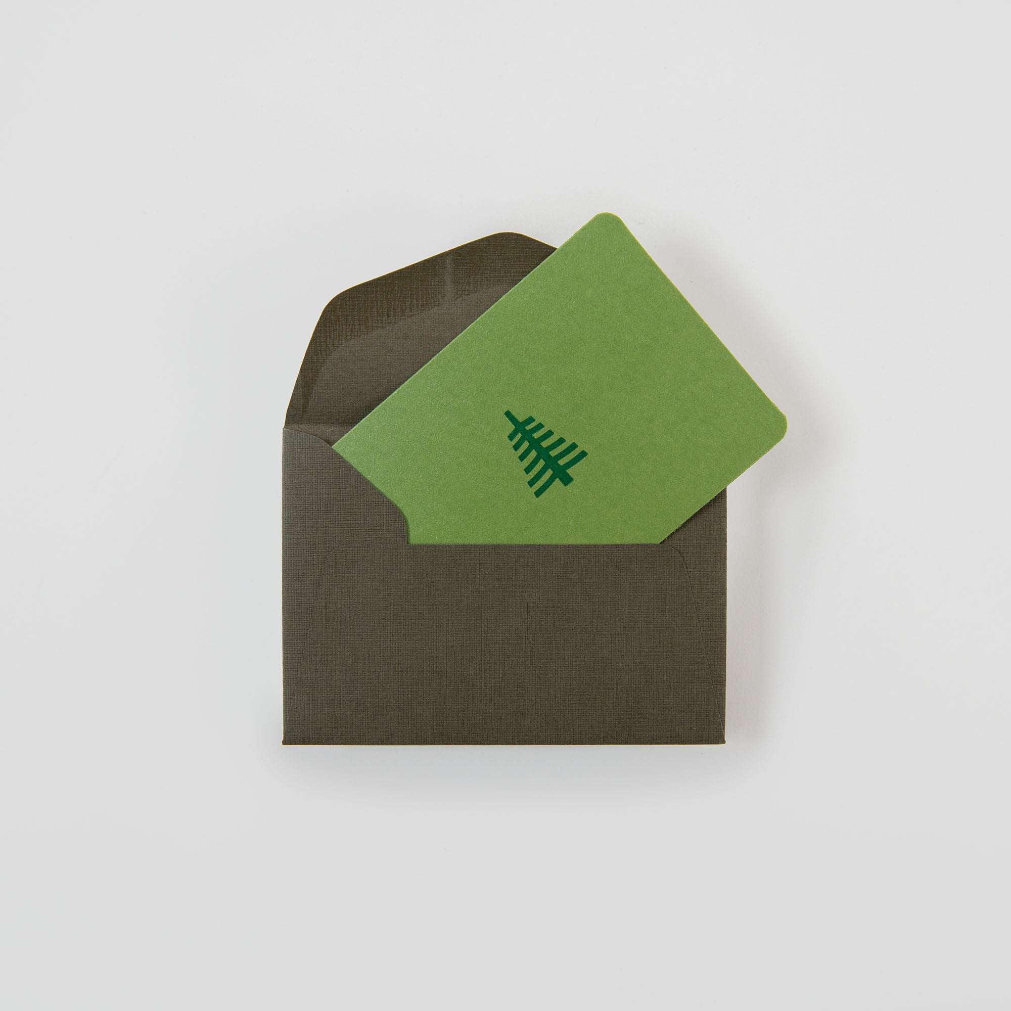 Die "Tannenbaum groß - Mini Karte" von Le Typographe mit einem schlichten Baummotiv in Grün lugt aus einem dunkelbraunen Umschlag auf weißem Hintergrund hervor - perfekt für einen herzlichen vorweihnachtlichen Gruß.