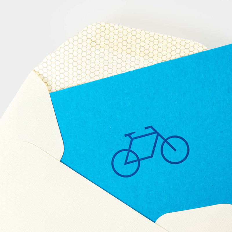 Ein weißer Umschlag mit Wabenmuster auf der Innenseite offenbart eine coole blaue Karte mit einem dunkelblauen Fahrradsymbol. Produkt: Fahrrad - Minikarte von Le Typographe.