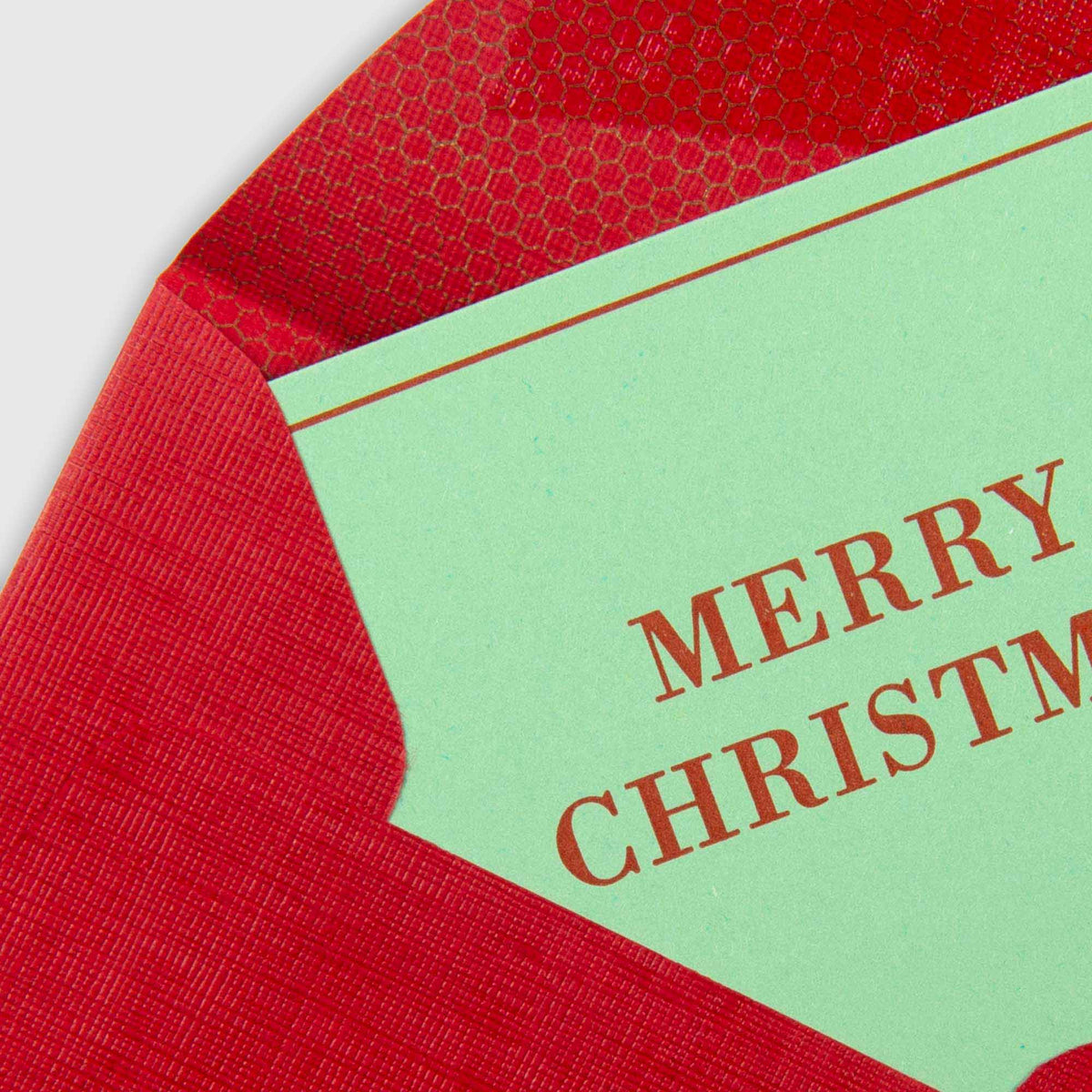 Eine minimalistische, hellgrüne Le Typographe Merry Christmas – Mini Karte-Karte im Letterpress-Stil mit braunem Text, die teilweise in einen roten Umschlag gesteckt ist.
