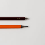 Eine Nahaufnahme von zwei Stiften auf weißem Hintergrund. Der obere Stift, „Mechanical Pencil – mit der Biene“ von Le Typographe, hat eine Kappe und ist dunkel mit einem kleinen gelben Logo. Der untere Stift hat keine Kappe, ist leuchtend orange und zeigt nach rechts.