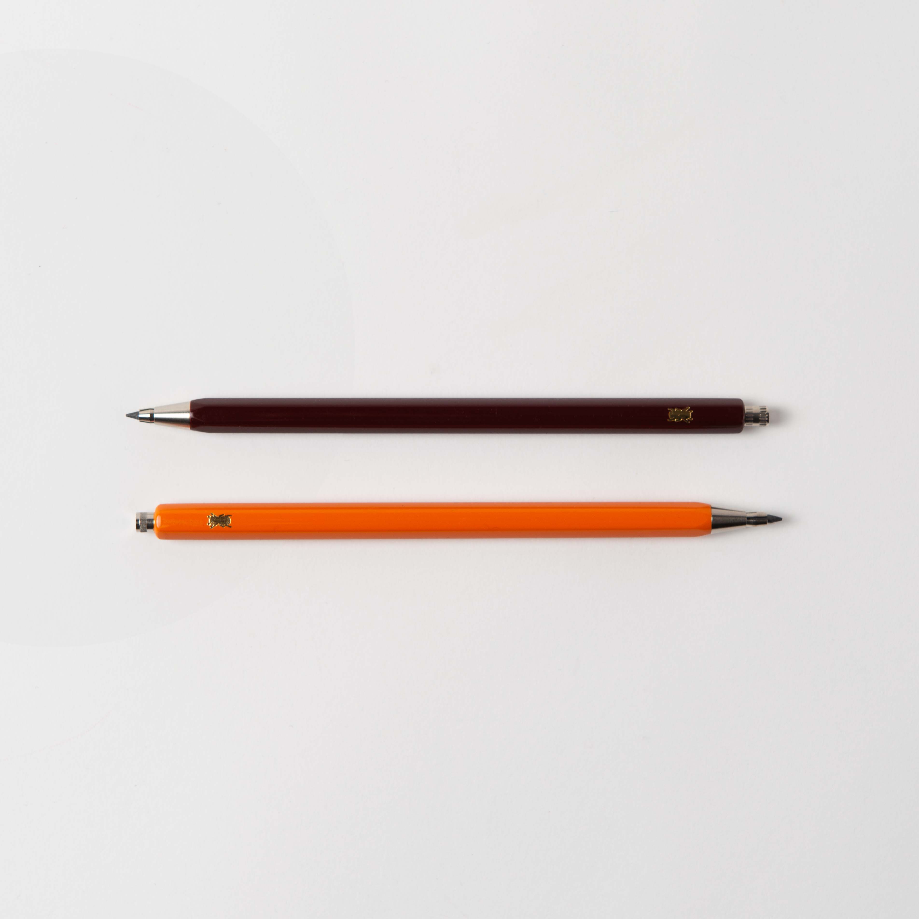Zwei „Mechanical Pencil – mit der Biene“ von Le Typographe liegen parallel auf einer weißen Fläche. Der obere Bleistift ist dunkelbraun, der untere leuchtend orange. Beide haben glatte silberne Spitzen und kleine Metallkappen, perfekt zum Skizzieren von Grußkarten- oder Grußkarten-Designs.