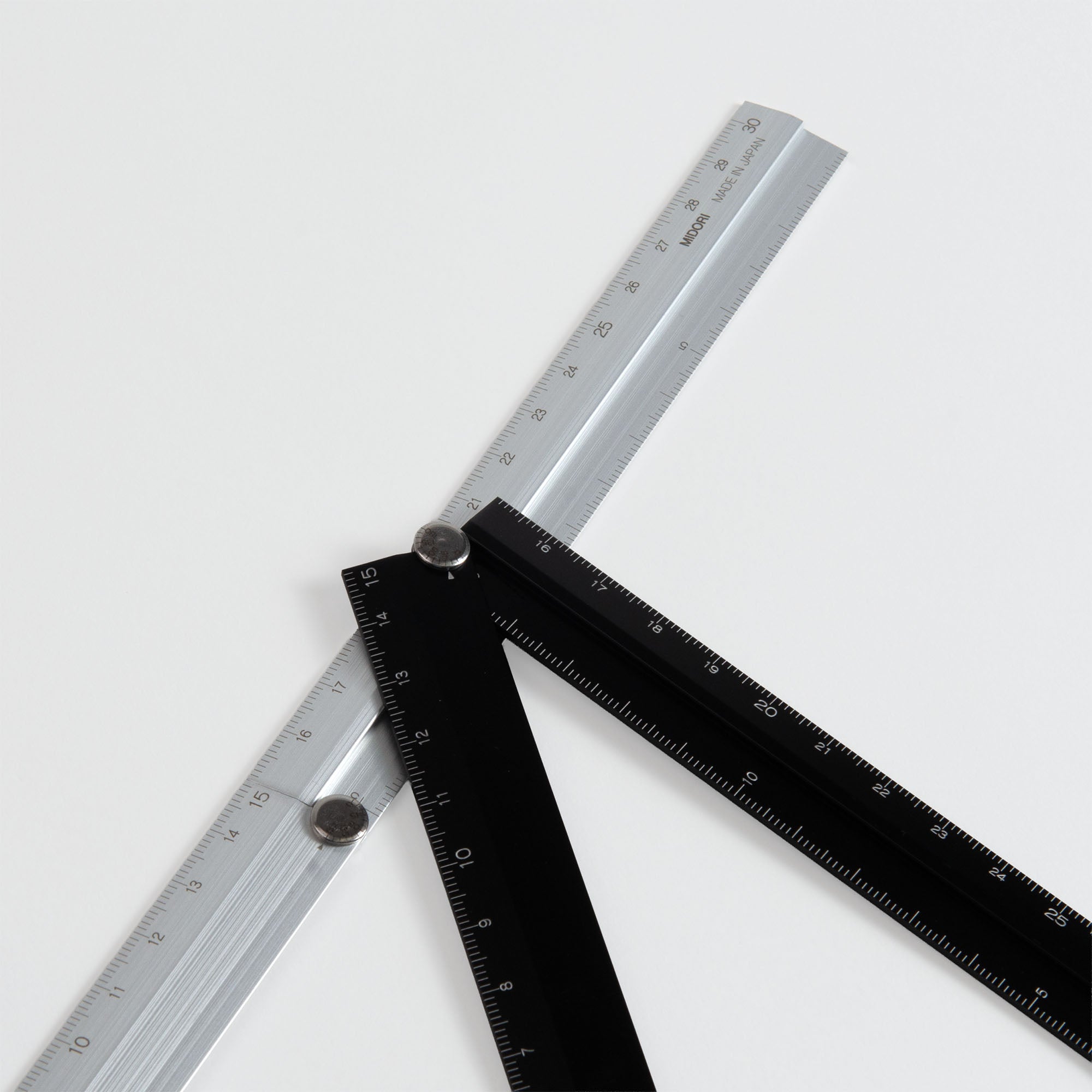 Drei Metalllineale - zwei schwarze und ein silbernes - sind an einem zentralen Drehpunkt verbunden und auf einem weißen Hintergrund ausgebreitet. Das Werkzeug, das Midori Klapp-Lineal von Midori, ähnelt einem geometrischen Messinstrument.