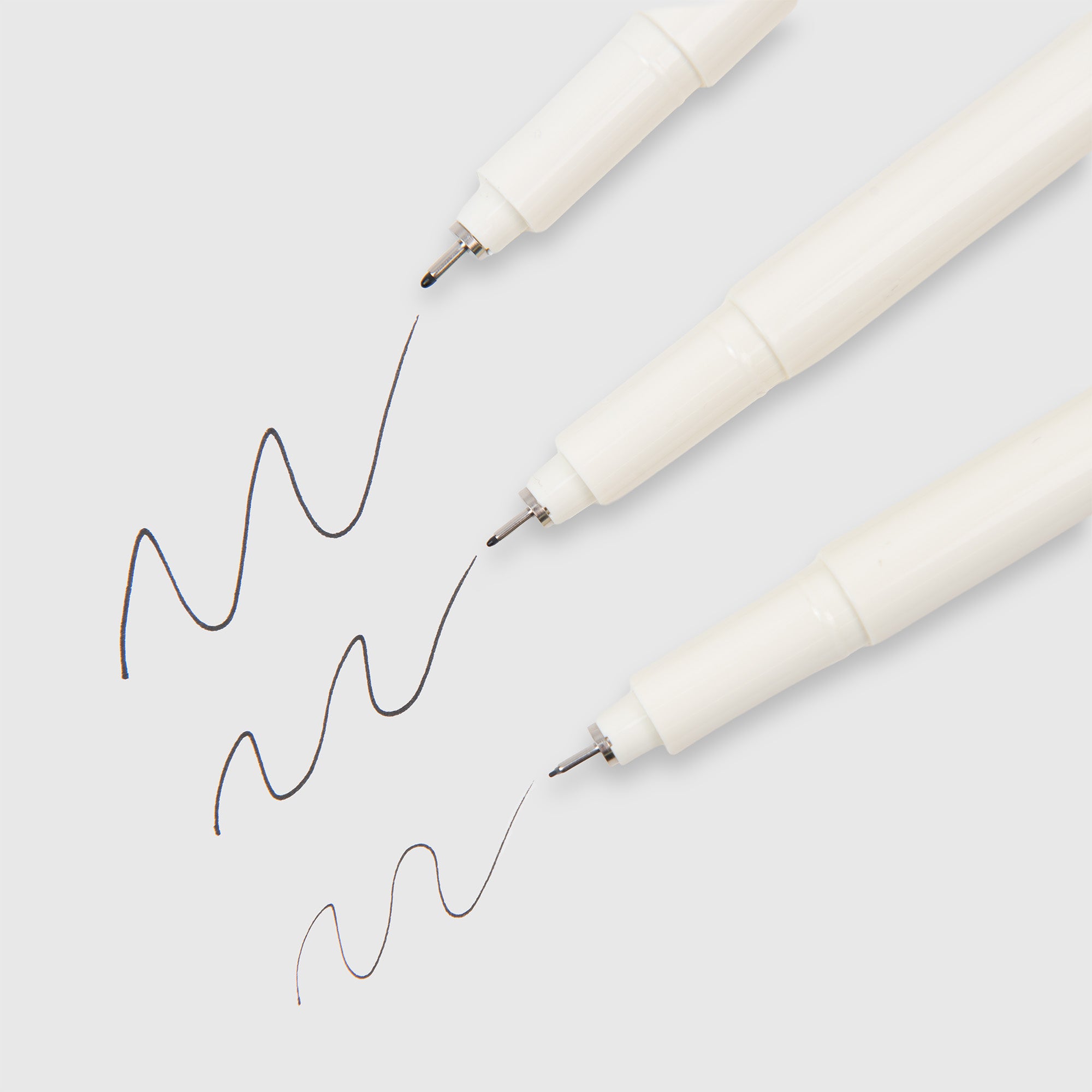 Drei weiße Midori MD Drawing Pens mit jeweils unterschiedlicher Spitzengröße zeichnen wellenförmige schwarze Linien auf hellem Hintergrund, um den Kontrast in der Strichstärke hervorzuheben. Diese Stifte von Midori verwenden wasserbasierte Pigmenttinte für gleichmäßige, konsistente Ergebnisse.