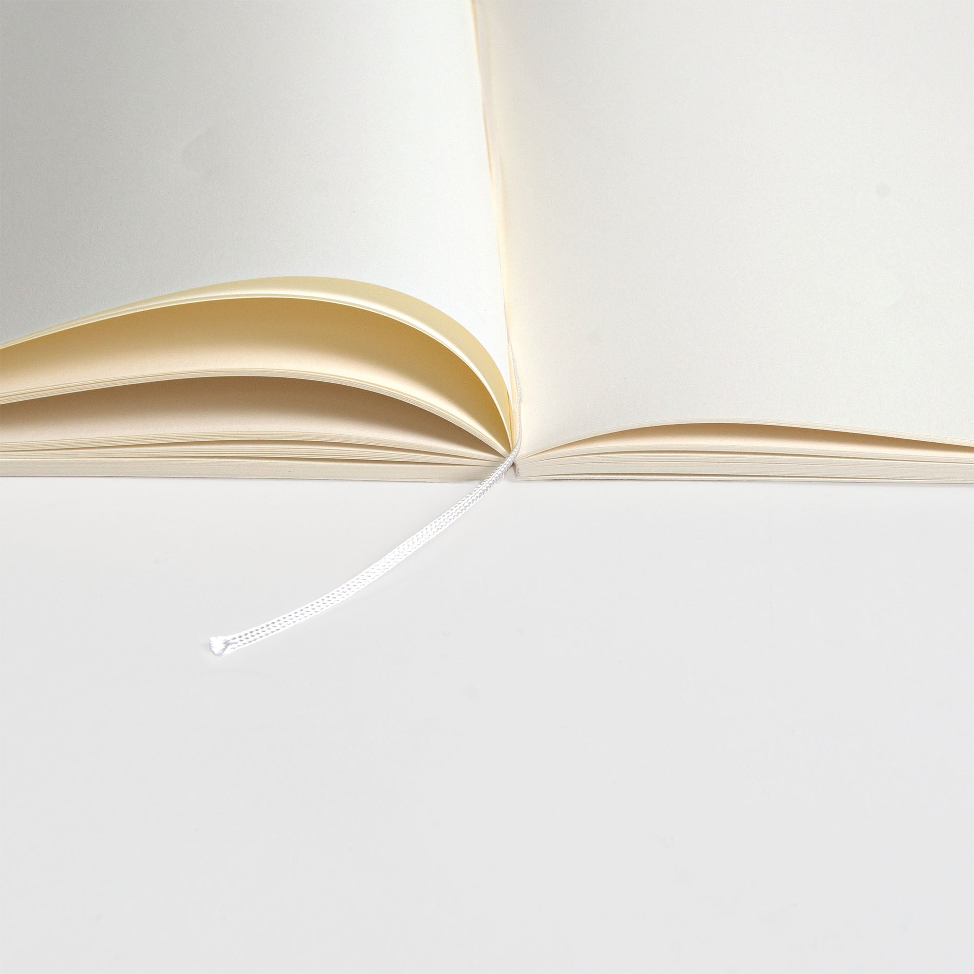Ein aufgeschlagenes Midori MD Notizbuch A5 Baumwolle liegt flach auf einer weißen Oberfläche und zeigt seine cremefarbenen Seiten aus MD Baumwollpapier und das Lesezeichen mit weißem Band. Die Fadenheftung bietet eine lange Haltbarkeit.