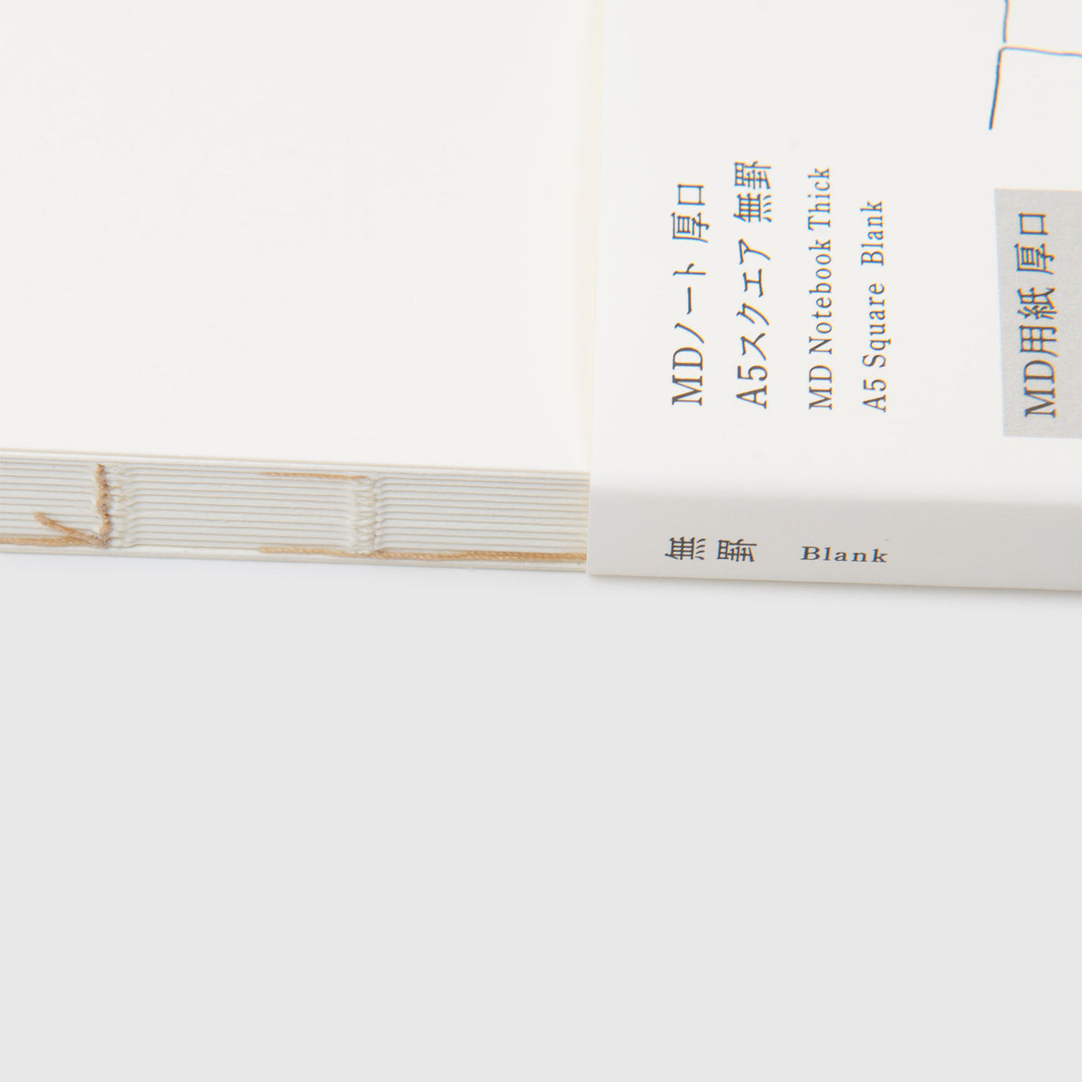 Nahaufnahme eines aufgeschlagenen, leeren Midori MD-Notizbuchs „Thick“ – A5, quadratisch, blanko, mit sichtbarer Heftung am Buchrücken. Auf dem Einband befindet sich sowohl japanischer als auch englischer Text, der es als Midori MD-Notizbuch im A5-Format, dicke Ausführung, mit leeren Seiten ausweist. Dieses von Midori aus MD Paper Thick 141 g/m² hergestellte Notizbuch vermittelt ein hochwertiges Gefühl – ideal zum Skizzieren oder Schreiben.