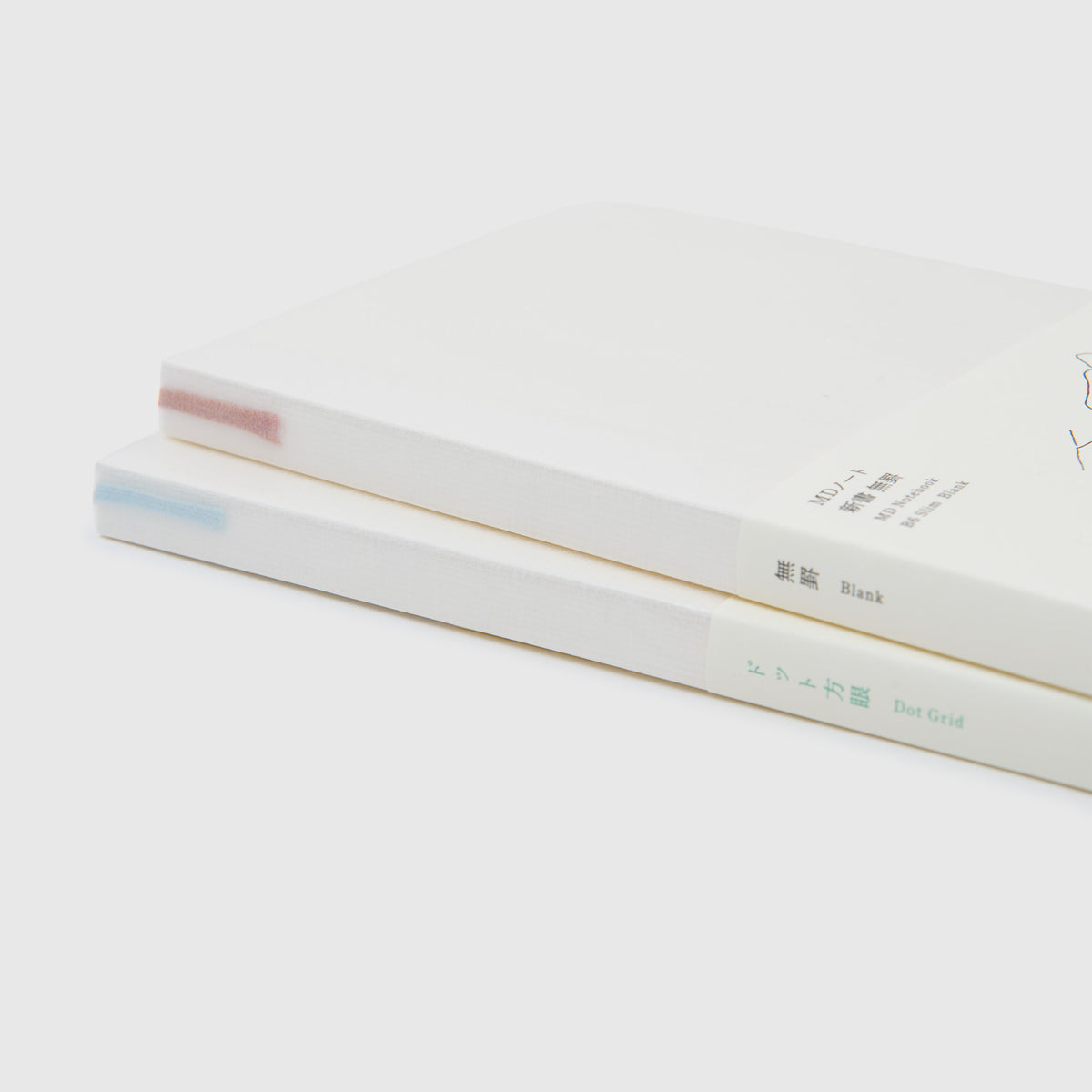 Zwei minimalistische weiße Midori MD-Notizbücher – B6 Slim – sind übereinander gestapelt; eines hat eine blaue Markierung auf dem Buchrücken mit der Aufschrift „Dot Grid“, das andere eine rote Markierung mit der Aufschrift „Blank“, beide mit japanischem und englischem Text auf dem Buchrücken. Diese Midori-Notizbücher im perfekten B6-Slim-Format sind für Füllfederhalter geeignet – ideal für alle, die Wert auf geschmeidiges Schreiben und zurückhaltende Eleganz legen.