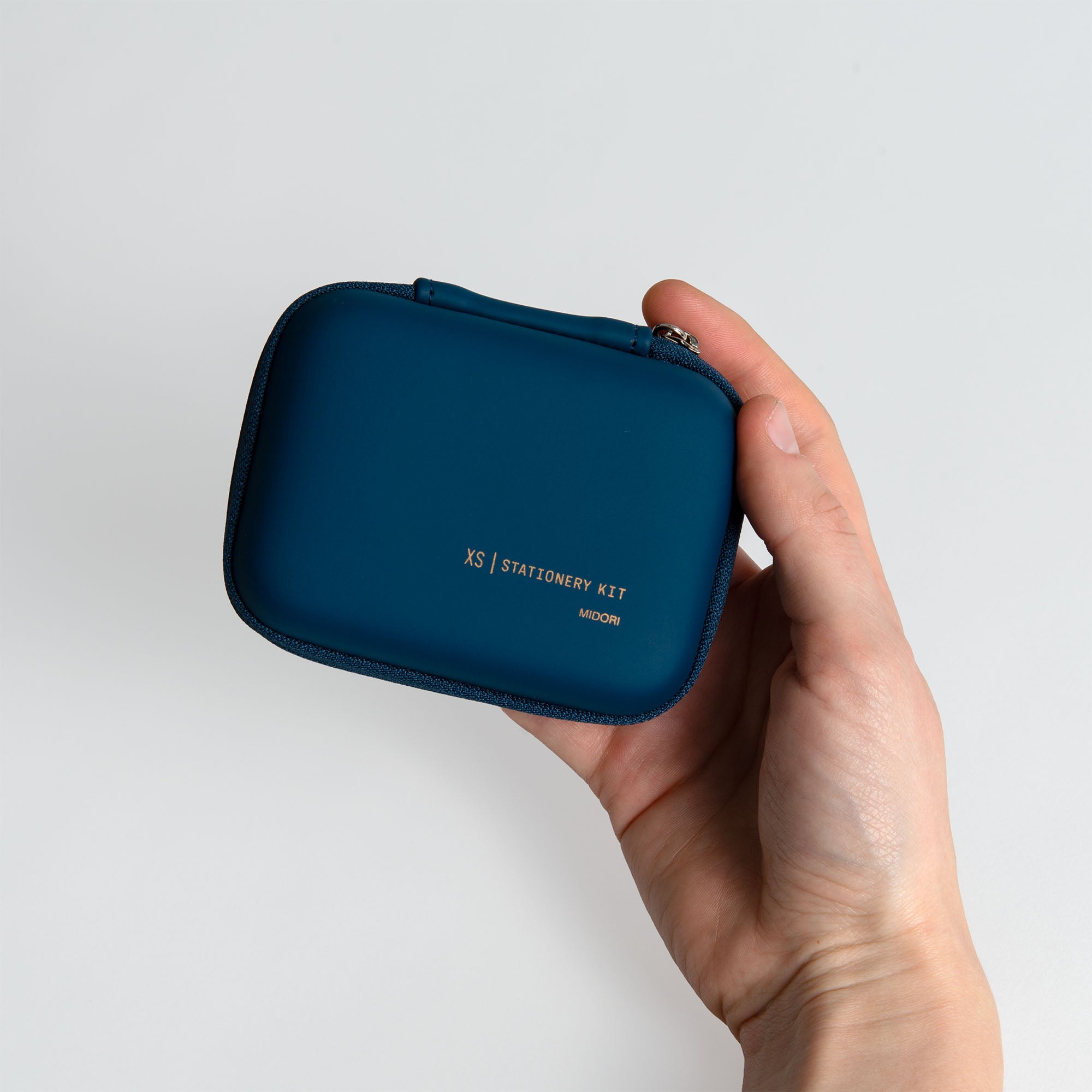 Eine Hand hält ein kompaktes, rechteckiges blaues Midori XS Stationery Kit mit Reißverschluss, beschriftet mit XS | STATIONERY KIT - ideal für ein tragbares Schreibtischset - vor einem schlichten weißen Hintergrund. Marke: Midori.
