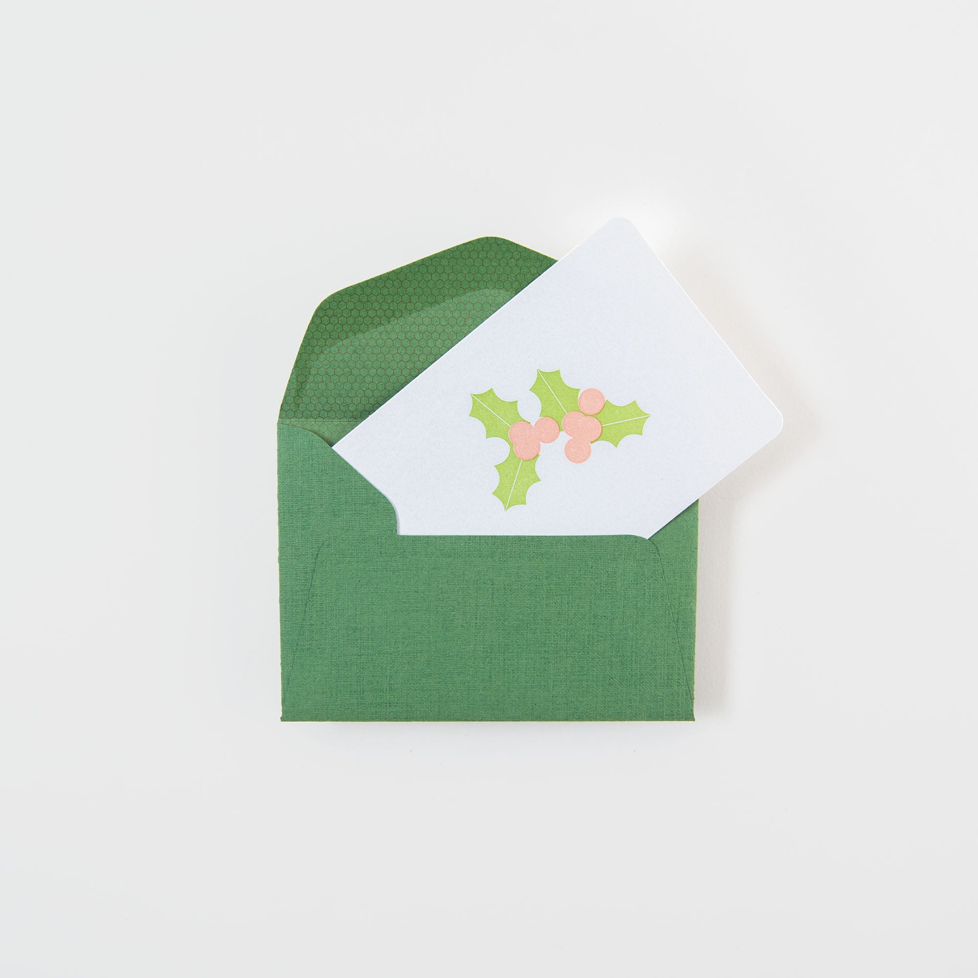 Ein geöffneter grüner Umschlag enthüllt die "Mistelzweig - Mini Karte" von Le Typographe, eine Weihnachtskarte mit Stechpalmenblättern und rosa Beeren auf Weiß. Die festliche Karte wird auf einer schlichten weißen Fläche präsentiert.
