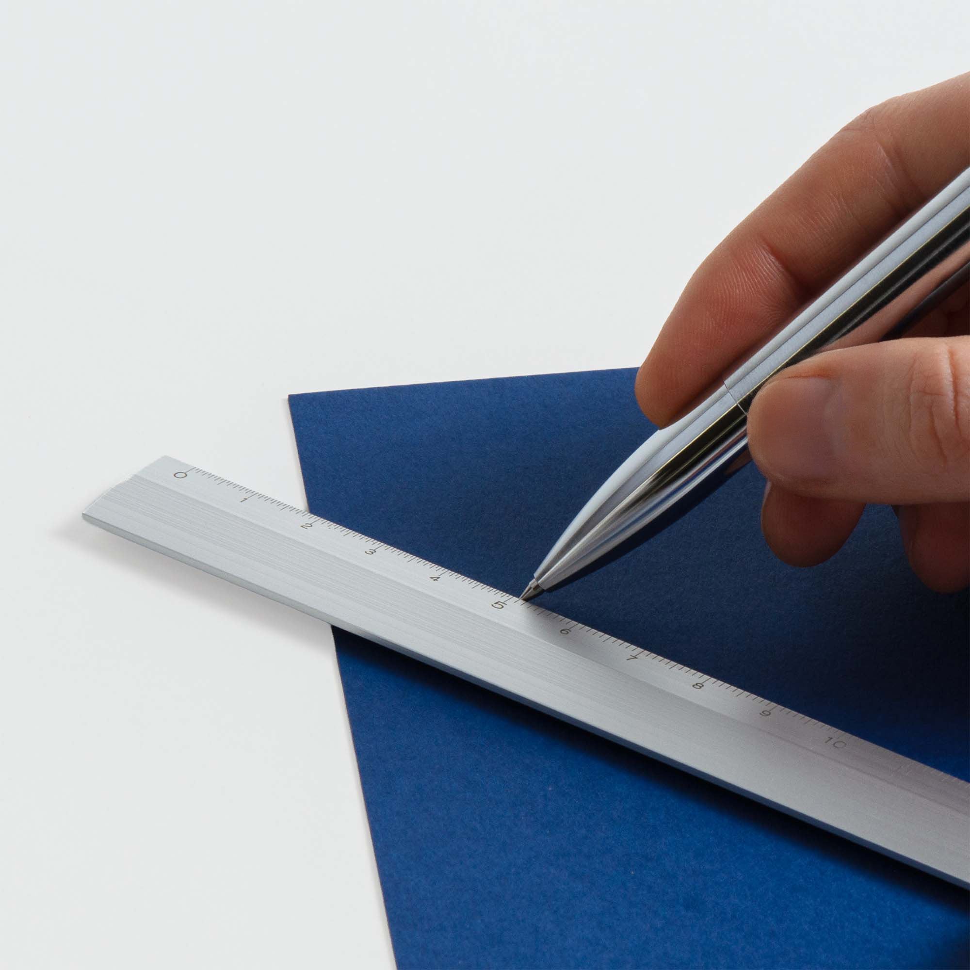 Eine Hand zeichnet mit dem silbernen Midori Klapp-Lineal von Midori eine gerade Linie auf ein blaues Blatt Papier vor einem weißen Hintergrund.