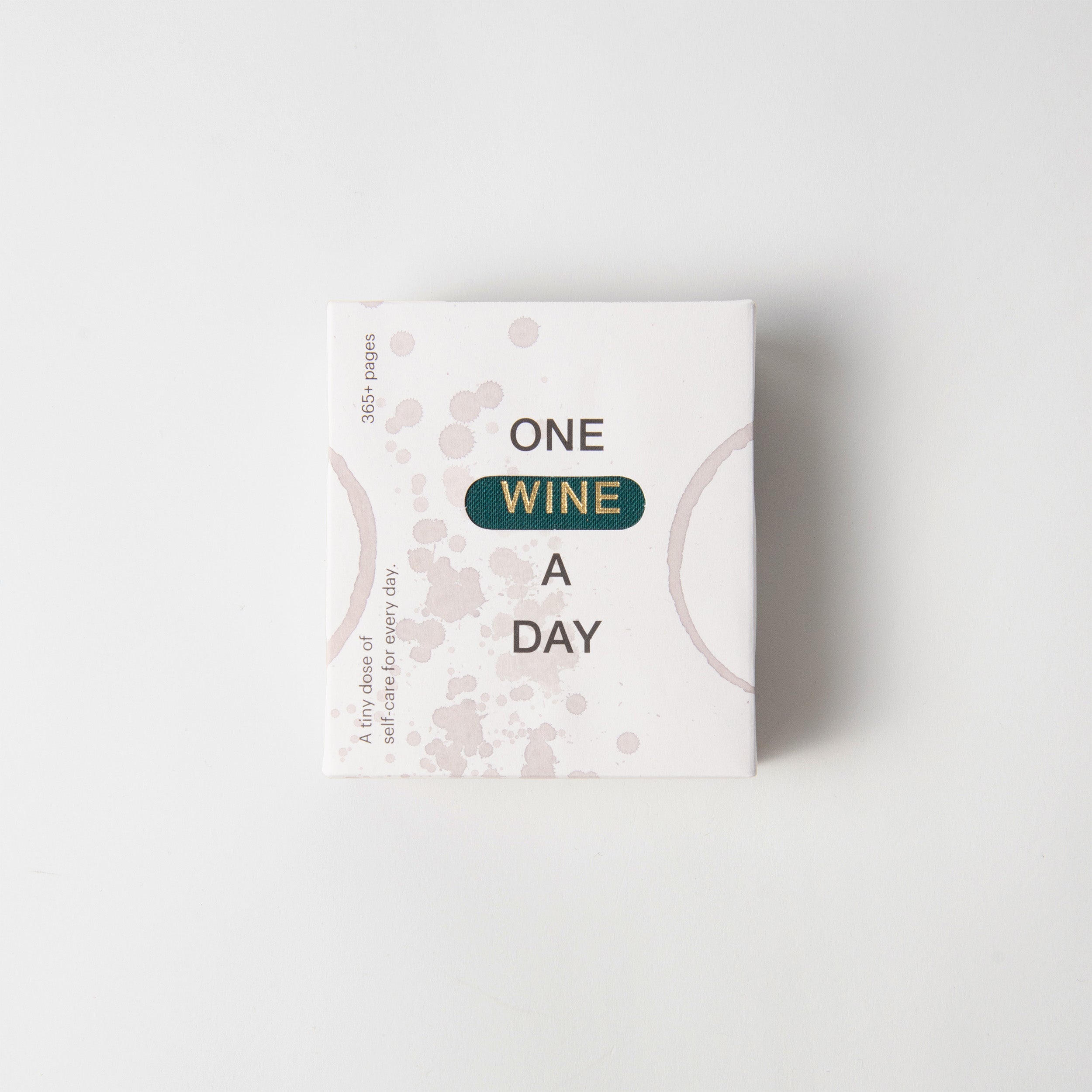 Auf einer quadratischen Box von paperlux.store mit minimalistischem Design steht in Fettschrift „One Wine a Day“, akzentuiert durch abstrakte rosa Flecken und geschwungene Linien, die an Weinspritzer auf weißem Hintergrund erinnern.