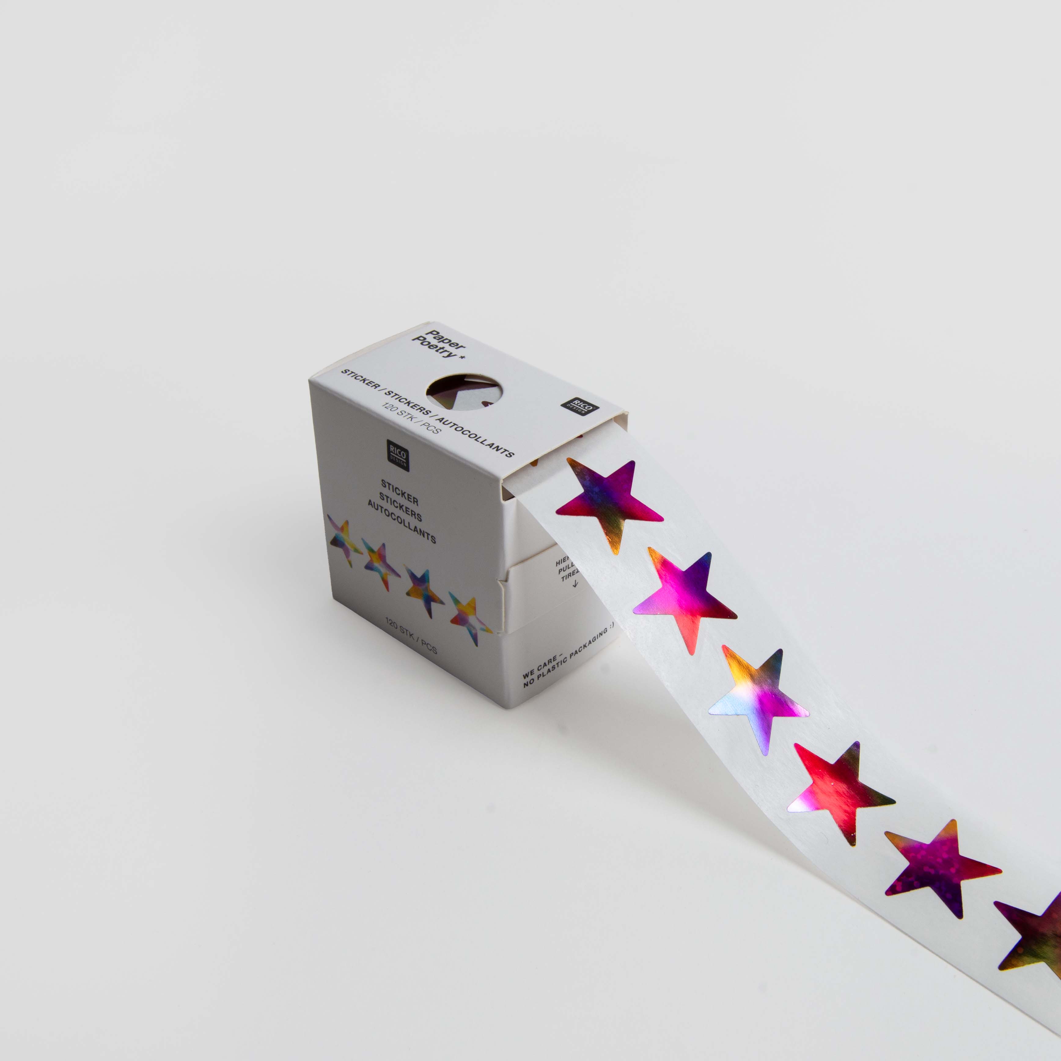 Eine weiße Paper Poetry Schachtel enthält eine Rolle glänzender, mehrfarbiger Paper Poetry Mini-Sticker Sterne Rainbow auf klarem Untergrund, auf dem mehrere Sterne aufgerollt sind. Der Hintergrund ist einfarbig weiß.