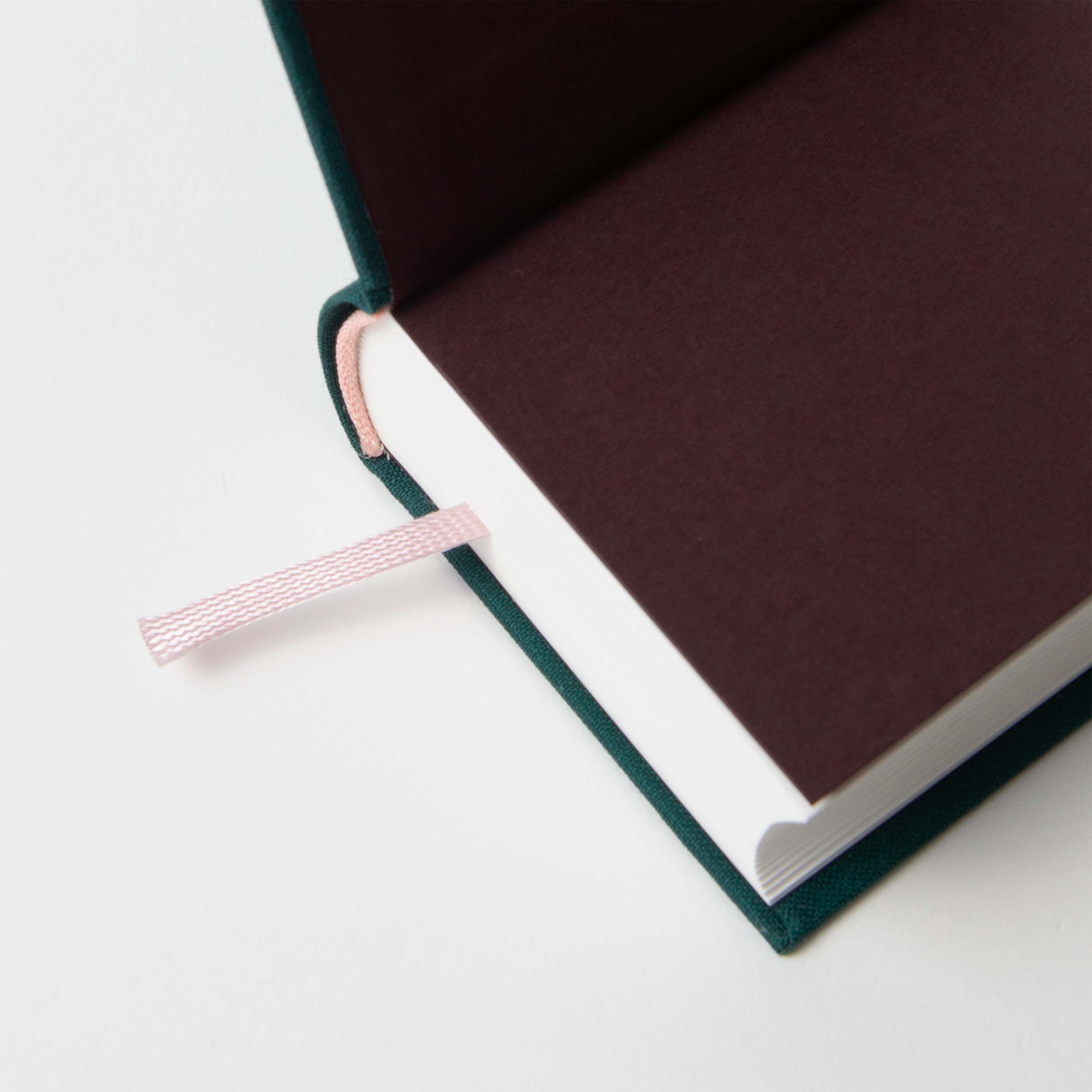 Das aufgeschlagene Hardcover-Journal One Wine a Day von paperlux mit dunkelbraunem Vorsatzpapier, weißen Seiten und einem zartrosa Lesezeichen liegt auf einer weißen Oberfläche. Dieses elegante Journal ist perfekt für Sommeliers und wird in Hamburg hergestellt.