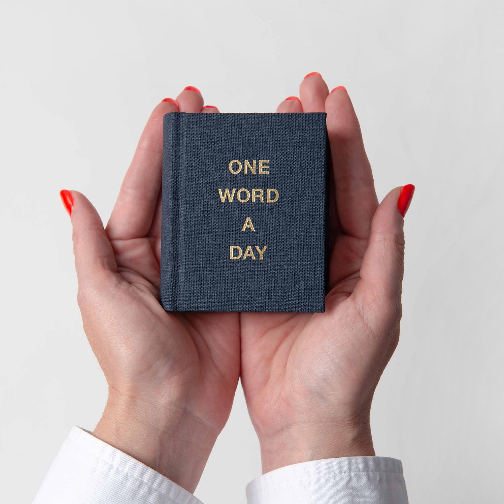 Eine Person mit rot lackierten Nägeln hält ein kleines OWAD-Klassiker-Notizbuch aus Papierlux mit der Aufschrift ONE WORD A DAY in goldenen Buchstaben vor einem schlichten weißen Hintergrund - perfekt als Geschenk.