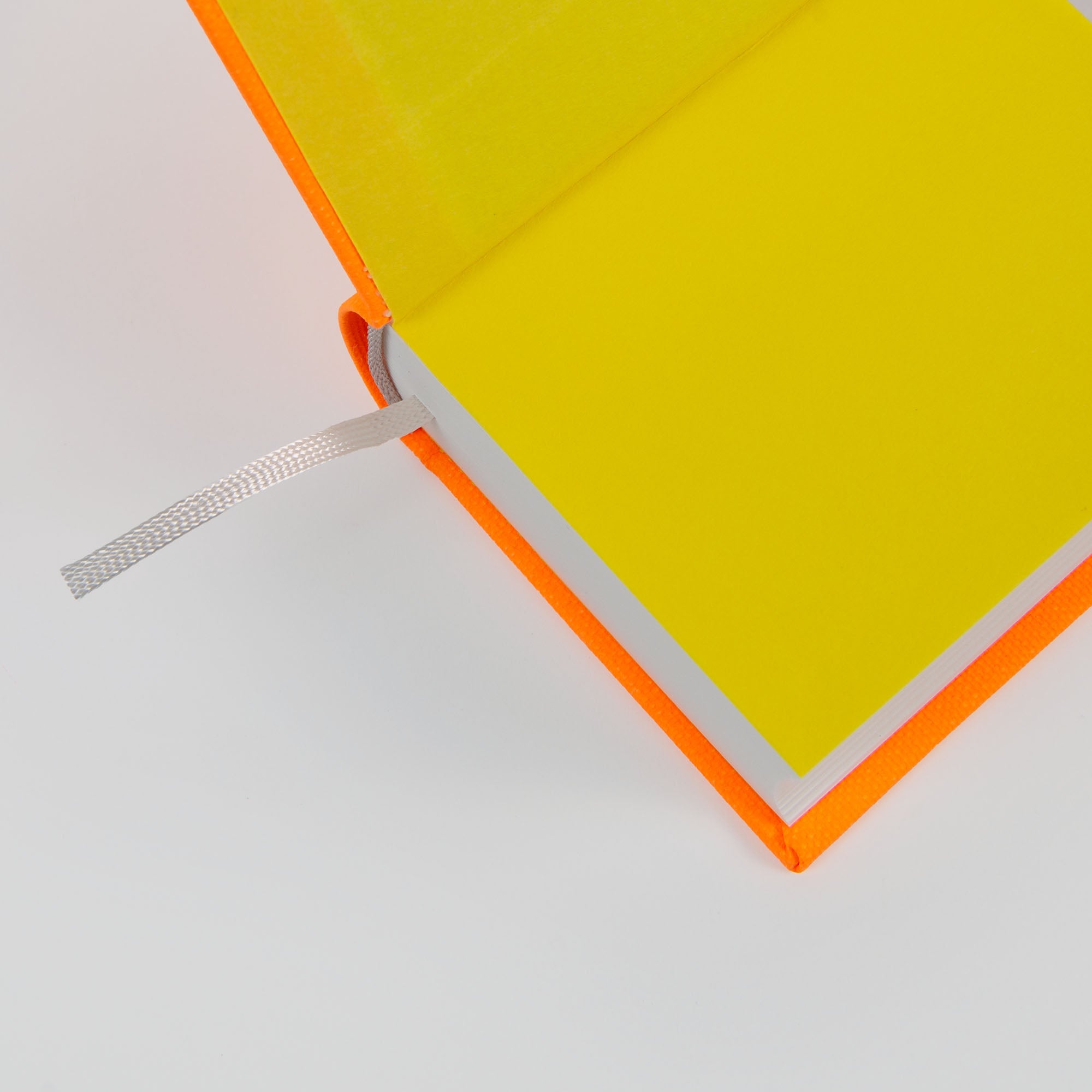 Das paperlux OWAD Neon ist ein aufgeschlagenes Notizbuch mit leuchtend gelben Seiten, einem orangefarbenen Einband und einem Lesezeichen mit silbernem Band - ideal für Tagebucheinträge oder die "One Word a Day"-Challenge. Eine durchdachte Geschenkidee, die vor einem weißen Hintergrund gezeigt wird.
