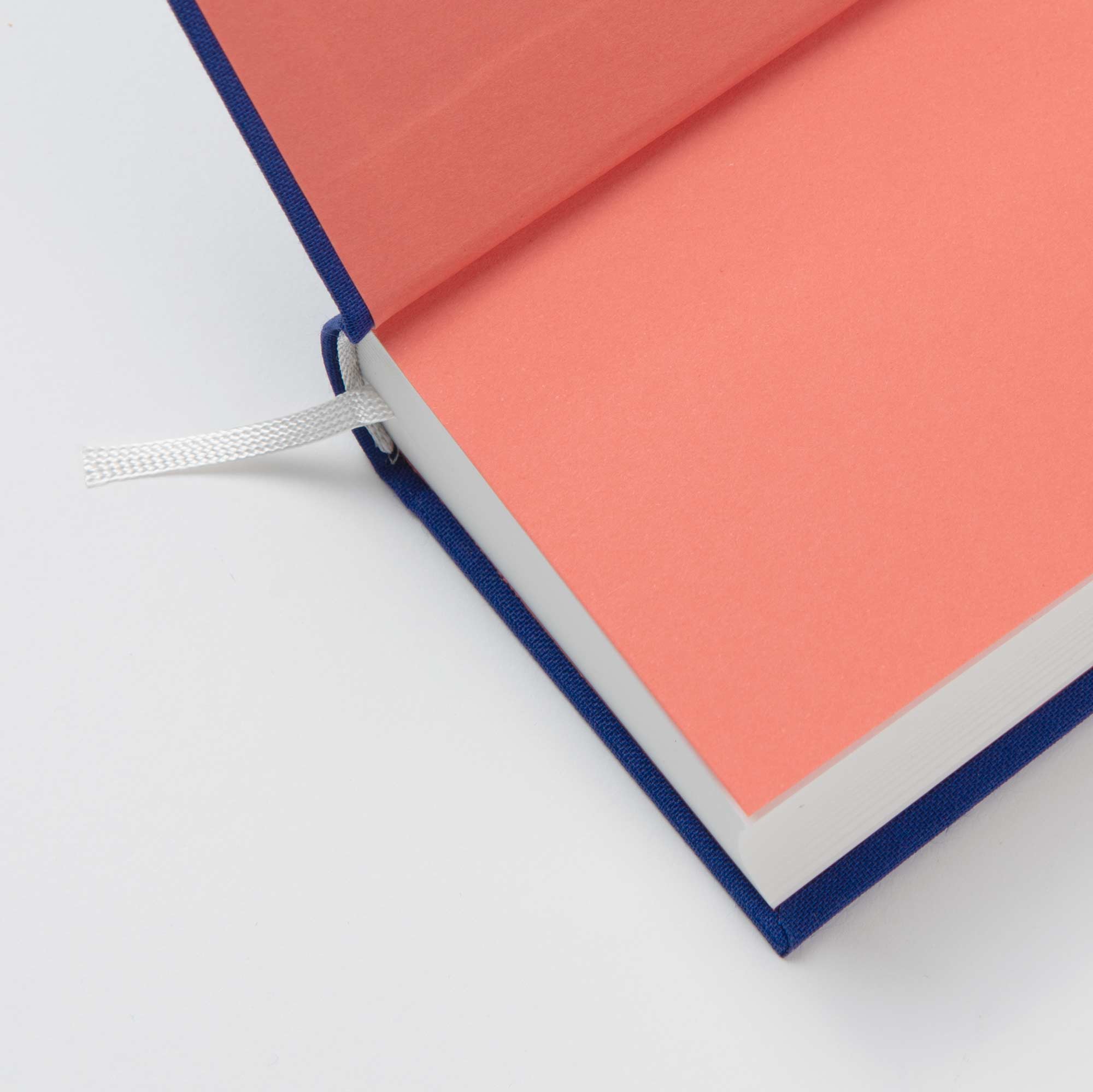 Das paperlux OWAD Shine Bright ist ein aufgeschlagenes Hardcover-Mini-Notizbuch mit blauem Einband, rosa Vorsatzpapier, weißen Seiten und einem Lesezeichen mit weißem Band, schräg von oben auf weißem Hintergrund betrachtet.