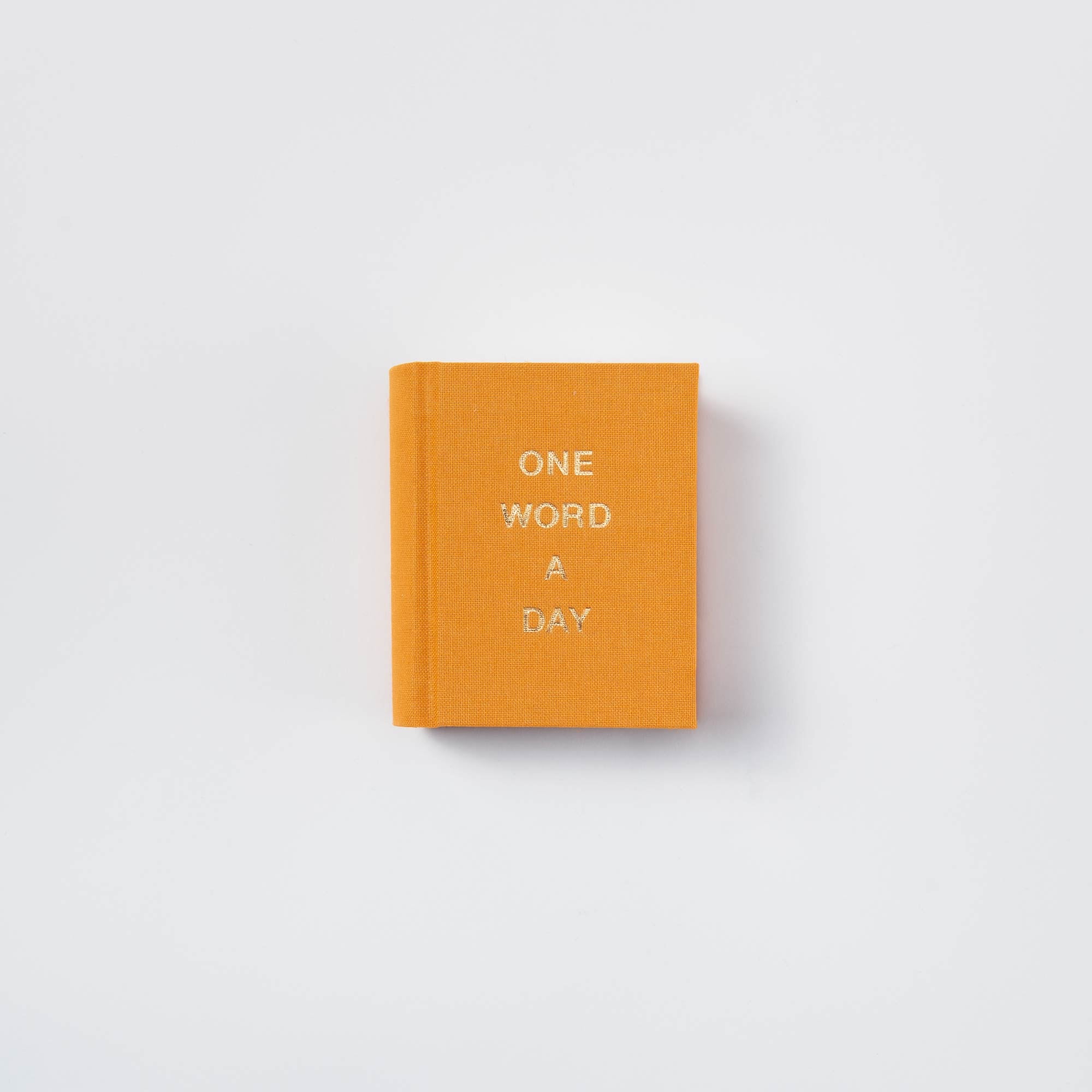 Ein leuchtend orangefarbenes paperlux OWAD Elements Leinenbuch mit "One Word A Day" in weißen Kappen, ideal als Mini-Tagebuch oder nachdenkliches Geschenk, zentriert auf einem sauberen weißen Hintergrund.