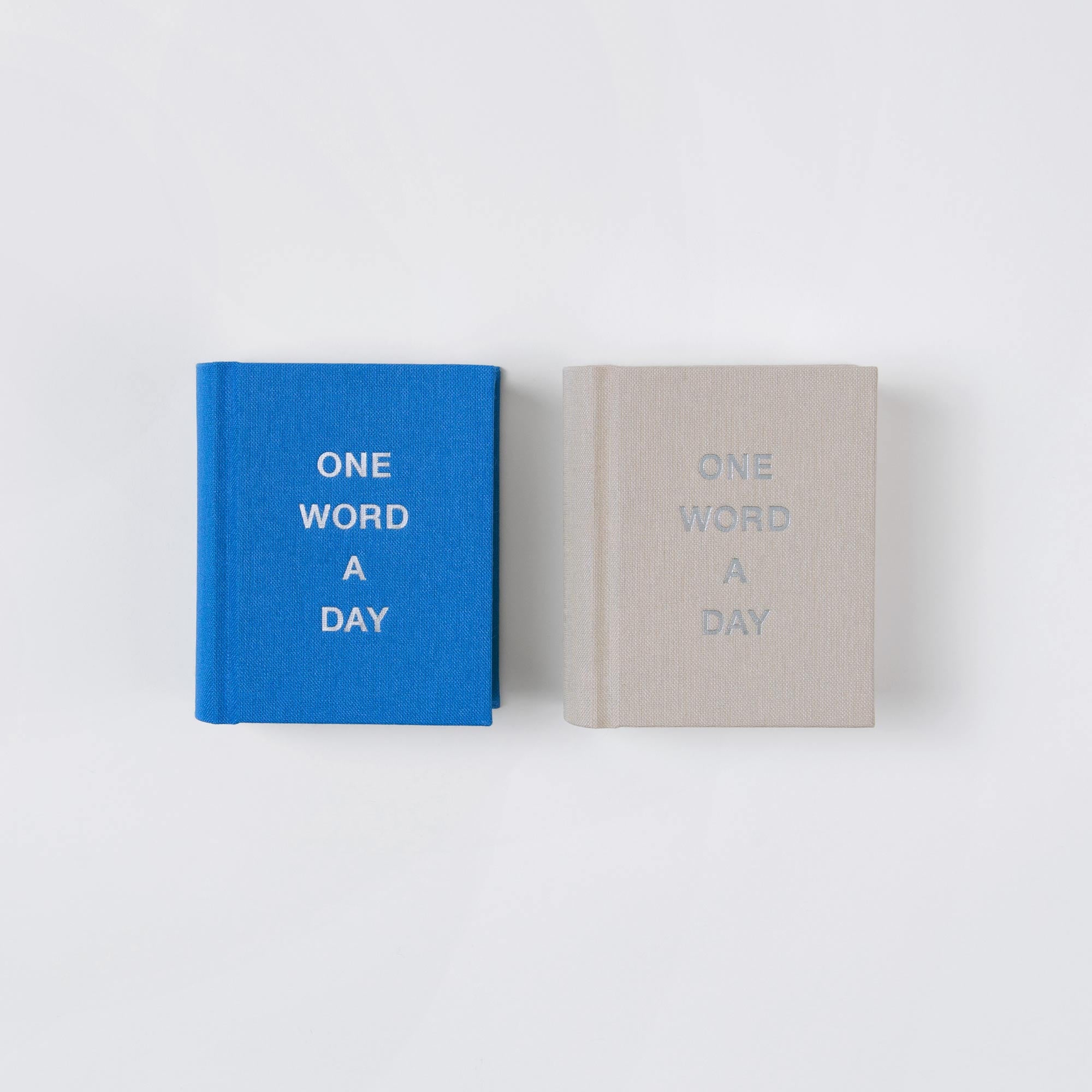 Zwei kleine fadengeheftete Leinenbüchlein von paperlux - eines blau, eines beige - liegen nebeneinander. Auf jedem OWAD Urban ist ONE WORD A DAY in Großbuchstaben eingeprägt, was sie zu einem charmanten Geschenk mit attraktiven Prägevarianten macht.