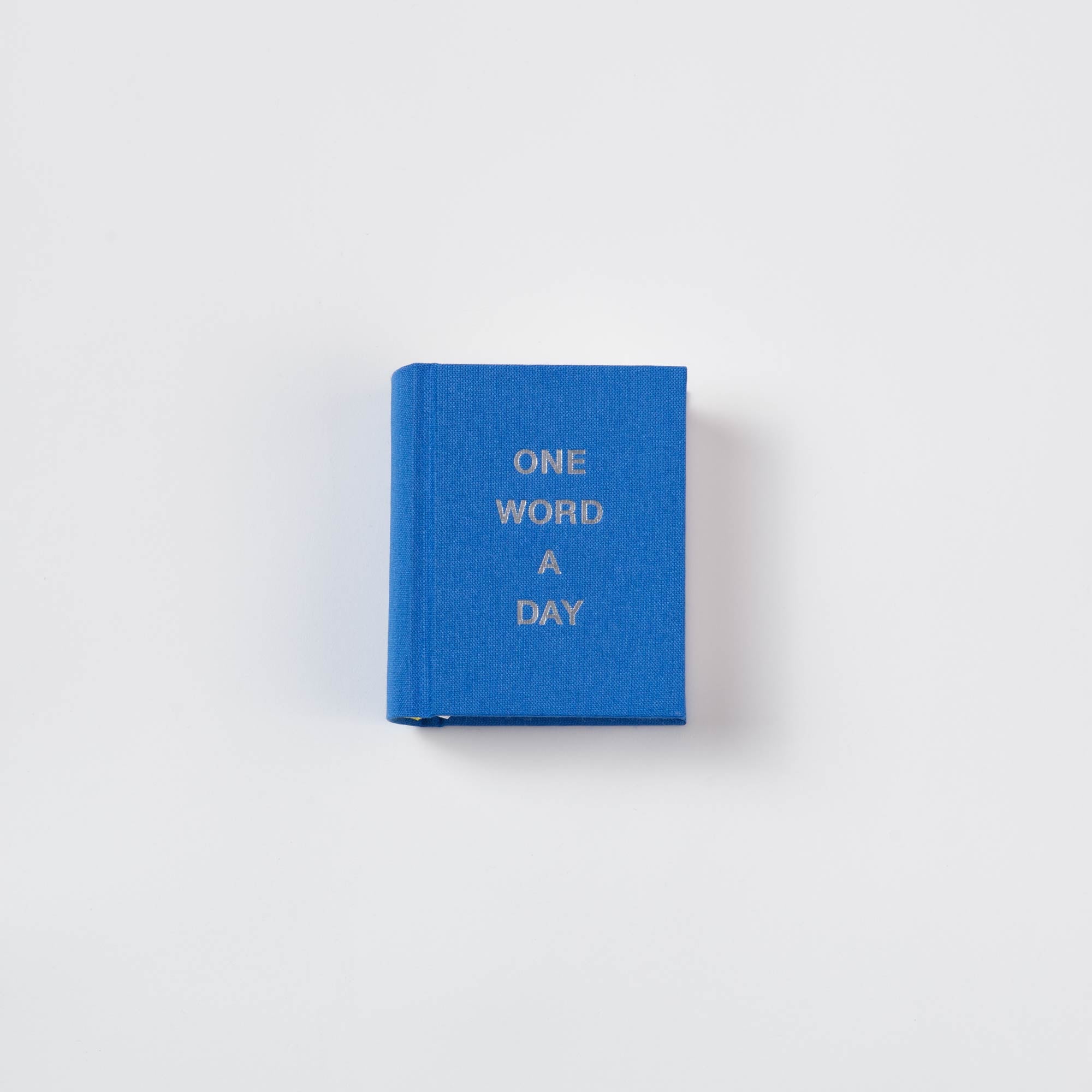 Ein kompaktes blaues OWAD Urban Leinen Notizbuch von paperlux, mit "ONE WORD A DAY" in Silber auf dem Cover, handgefertigt in Hamburg und fotografiert als charmantes Büchlein Geschenk vor einem schlichten weißen Hintergrund.