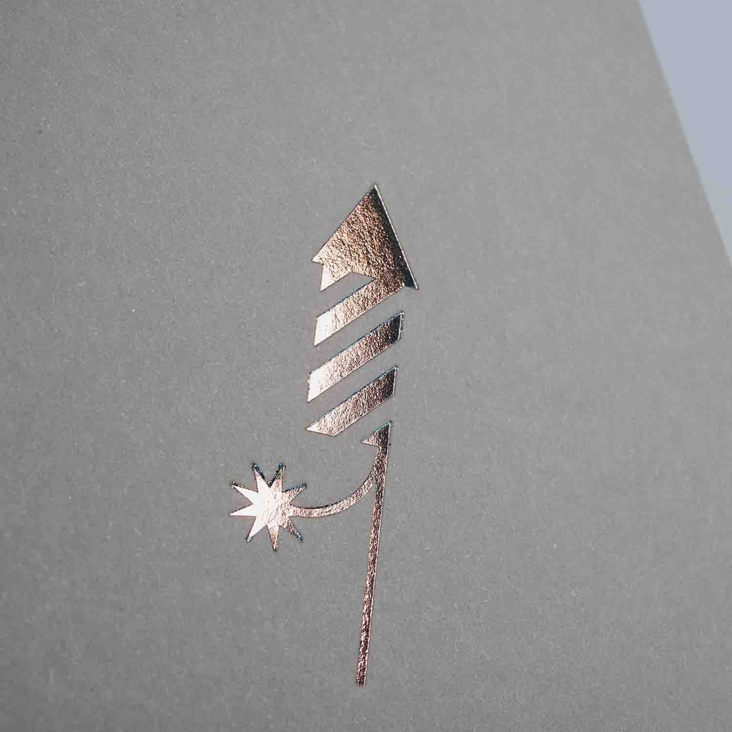 Die Karte Feuerwerk von paperlux.store zeigt ein Raketensymbol aus Goldfolie auf grauem Hintergrund und erinnert an eine Feuerwerksrakete mit einem Stern am unteren Rand. Diese minimalistische und elegante Neujahrsgruß-Klappkarte mit diagonalen Streifen auf dem Raketenkörper eignet sich perfekt für festliche Grüße.