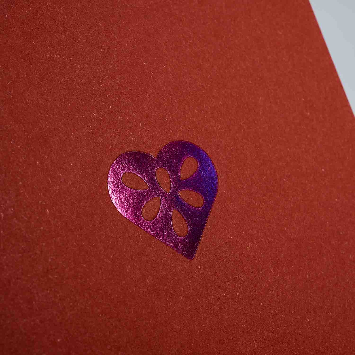 Ein metallisch glänzendes lilafarbenes Herz mit fünf Tränenausschnitten ziert einen strukturierten roten Hintergrund - ideal für festliche Momente mit dem Weihnachtskarten 3er-Set von paperlux.store.