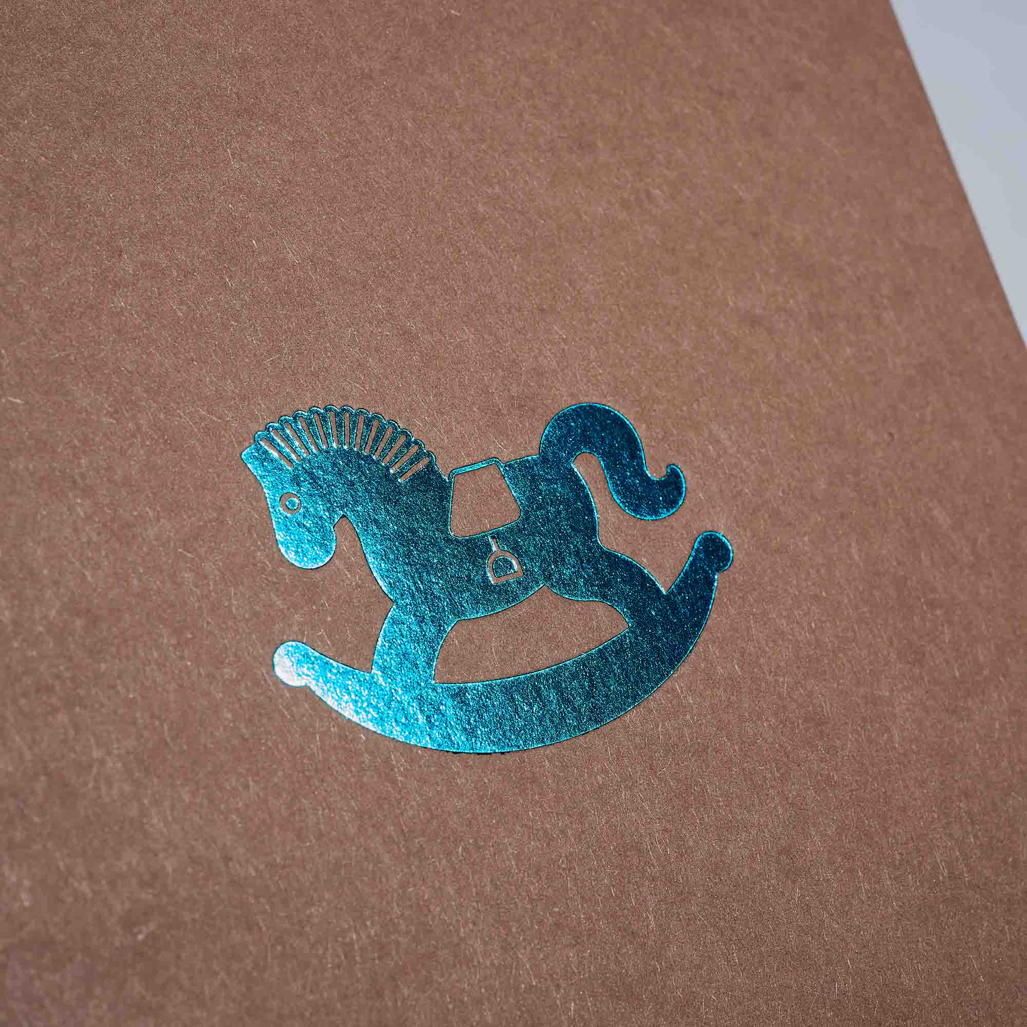 Die Karte Schaukelpferd von paperlux.store besticht durch eine braune Oberfläche mit einem glänzenden blaugrünen Folienstempel eines traditionellen Schaukelpferds mit Sattel und geschwungenen Kufen. Das nostalgische Design harmoniert mit dem strukturierten Hintergrund und eignet sich perfekt für eine elegante Geburtstags- oder Klappkarte.