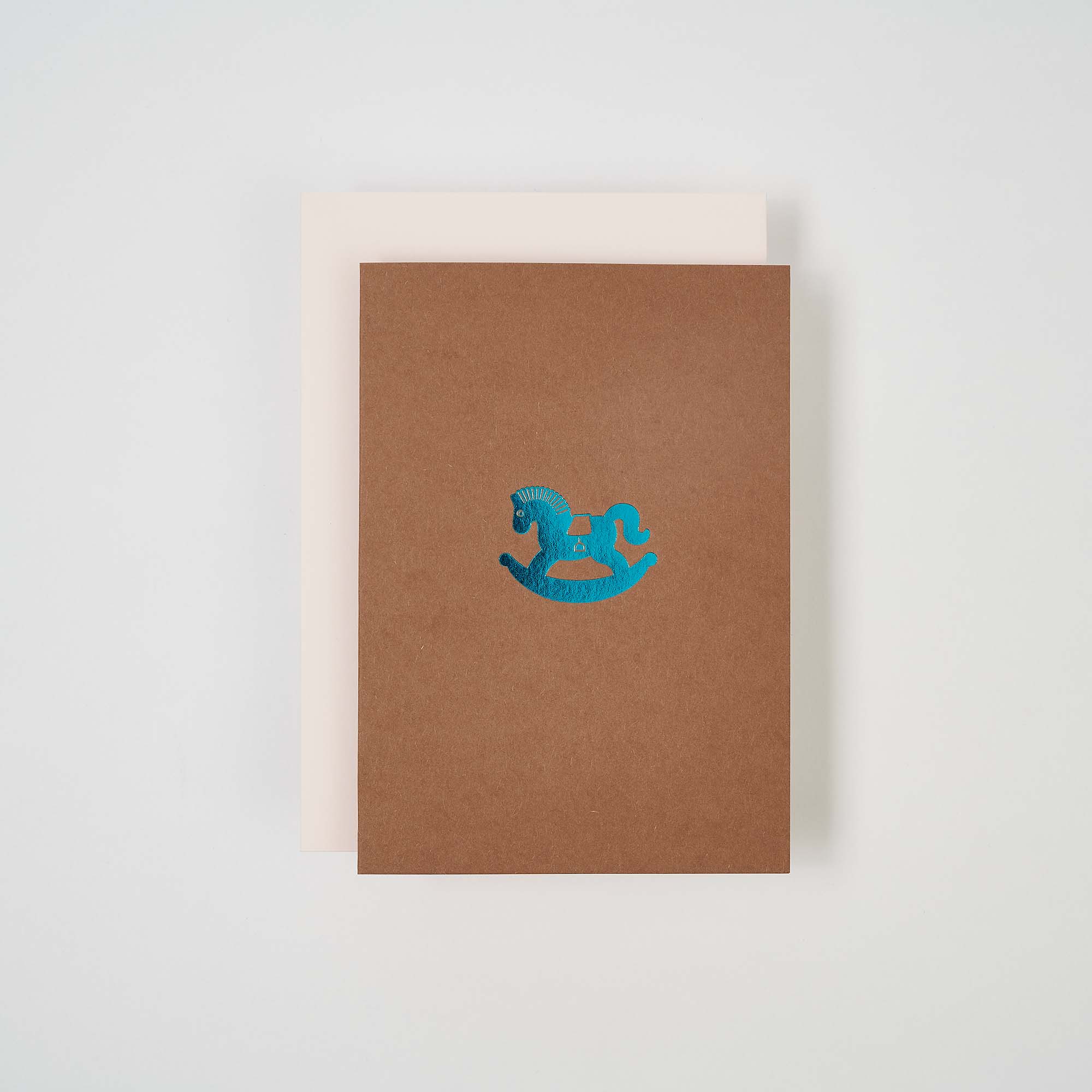 Die Karte Schaukelpferd von paperlux.store zeigt eine braune Geburtstagskarte mit einer blauen Schaukelpferd-Illustration auf einer weißen Klappkarte vor einem minimalistischen weißen Hintergrund.
