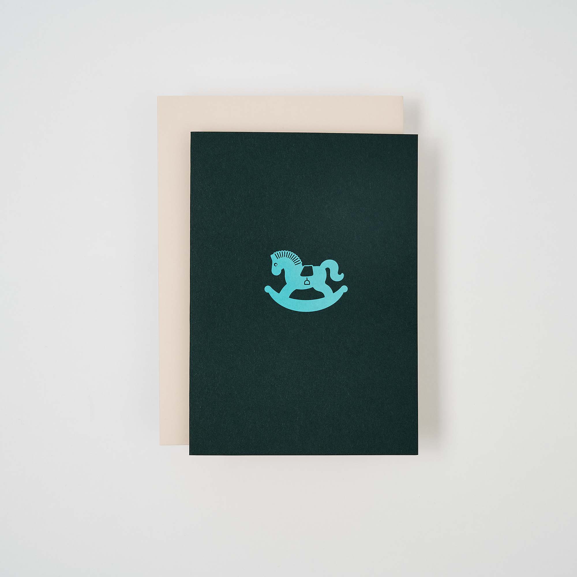 Die paperlux.store Karte Schaukelpferd besteht aus einer dunkelgrünen Klappkarte mit einem hellblauen Schaukelpferd-Symbol auf einer beigen Karte, das einen schönen Kontrast zum schlichten weißen Hintergrund bildet.