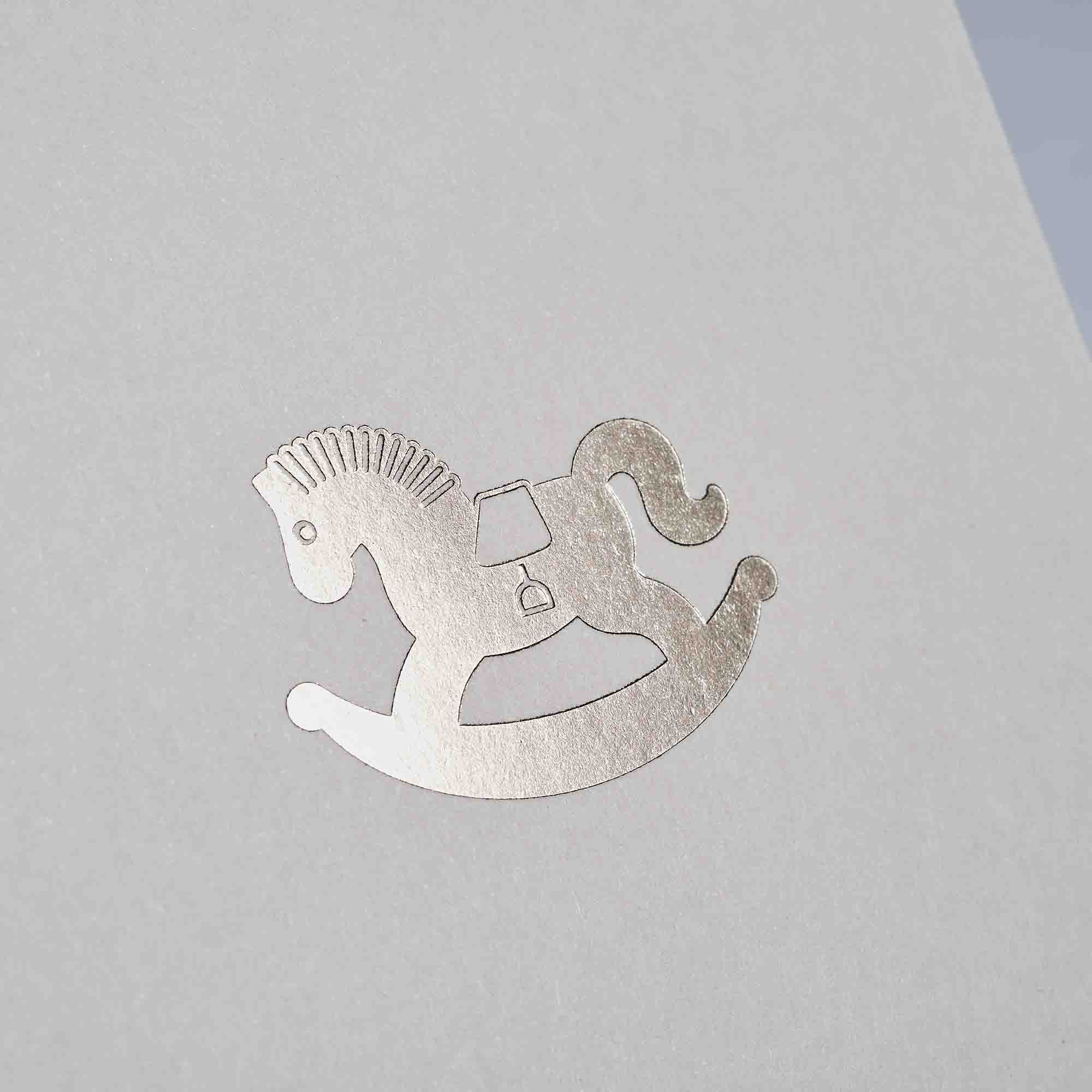 Die Karte Schaukelpferd von paperlux.store zeigt ein Schaukelpferd mit silberner Folienprägung, das elegant eine graue Oberfläche ziert.