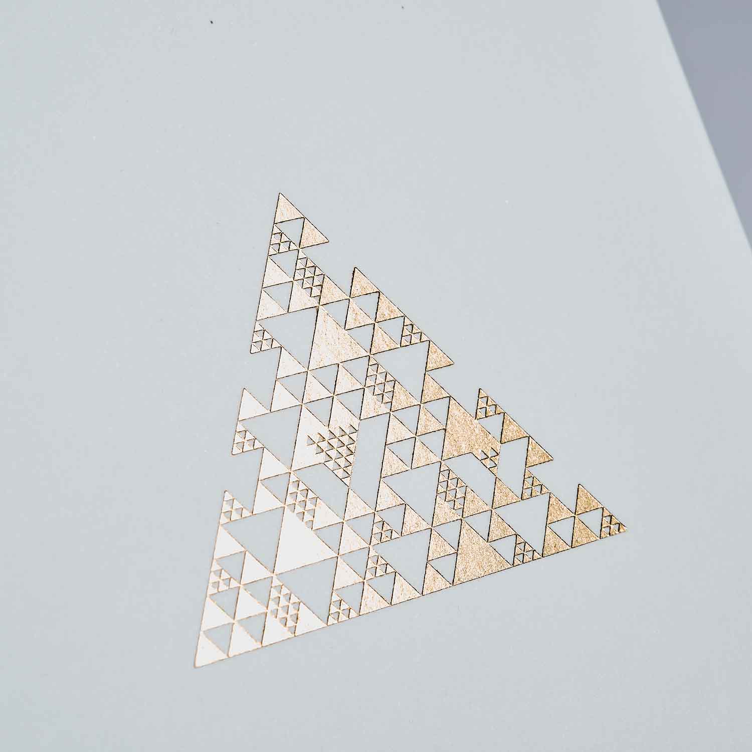 Ein goldenes geometrisches Dreieck aus einfarbigen und gemusterten Formen ist auf hellgrauem Untergrund zentriert, ideal für ein modernes Weihnachtskarten 2er-Set Snow & Coral von paperlux.store.