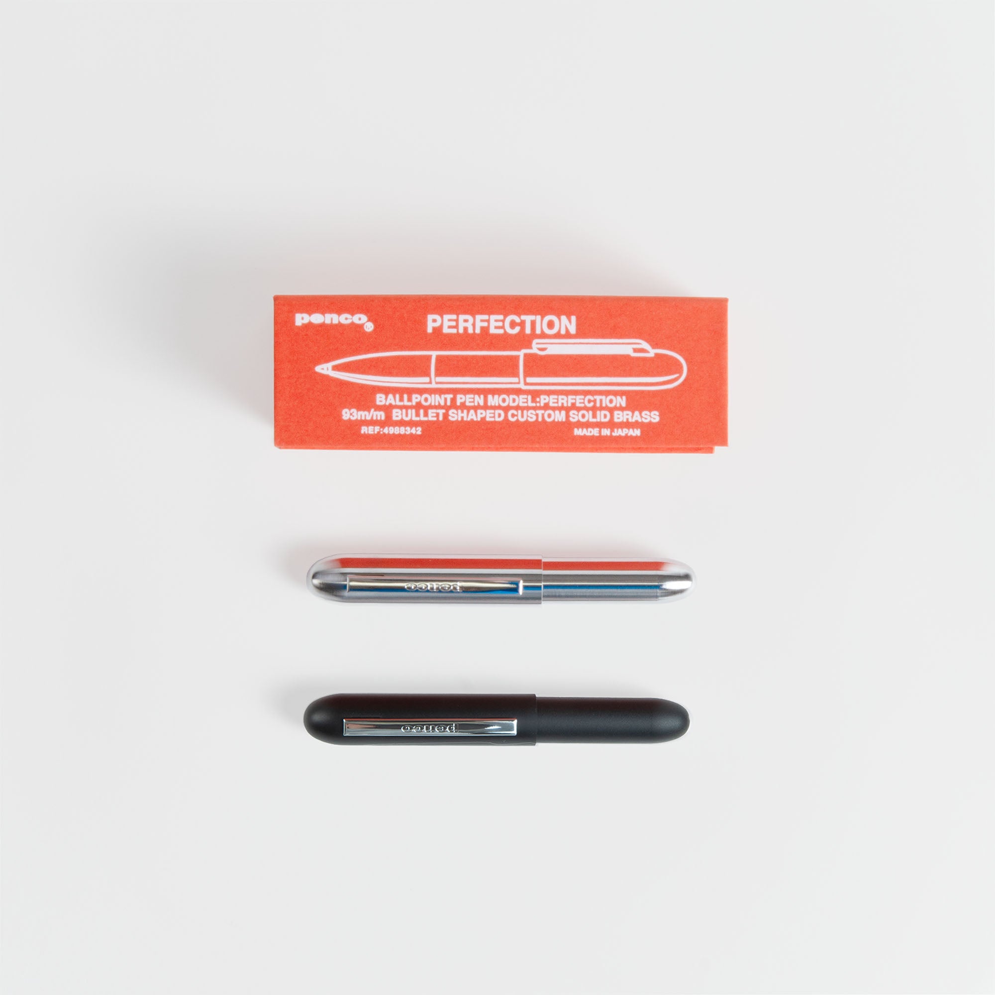 Eine rote Schachtel mit der Aufschrift PERFECTION ruht über einem paperlux.store Kompakt-Kugelschreiber - Bullet Pen mit schwarzem Messinggehäuse und schwarzer Kappe, alles auf einer weißen Fläche. Hergestellt in Japan.