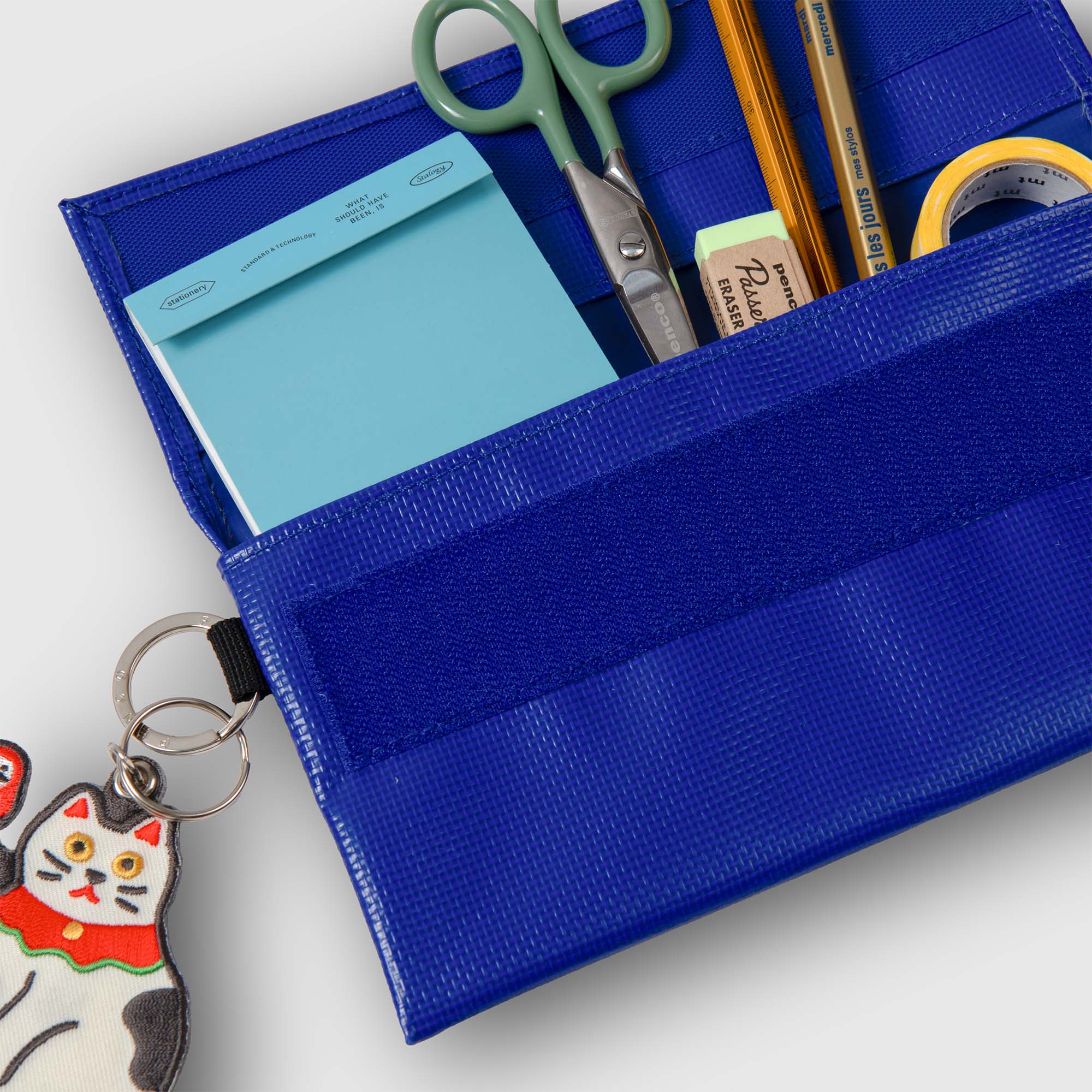 Nahaufnahme eines blauen Penco Carry Tite Tarp Pen Case aus wasserdichtem PVC-Planenmaterial, das Schere, Bleistift, Radiergummi, Klebeband und einen blauen Notizblock enthält. An der Federtasche ist ein Schlüsselanhänger mit einer Cartoon-Katze befestigt.