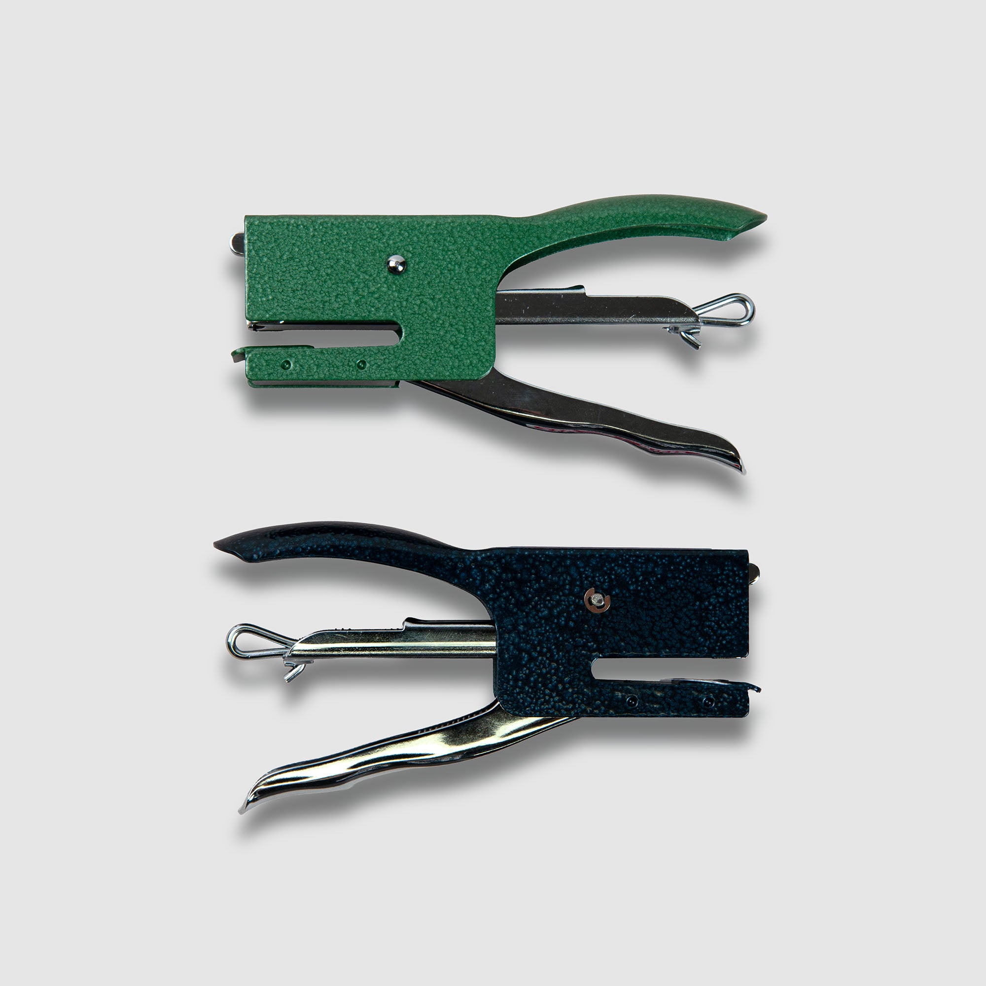Zwei Penco Hefter "Tacker", ein grüner und ein schwarzer, sind parallel auf einem einfarbigen hellgrauen Hintergrund angeordnet. Beide tragen die Marke Penco und verfügen über einen gebogenen Griff mit sichtbaren Metallklammermechanismen.