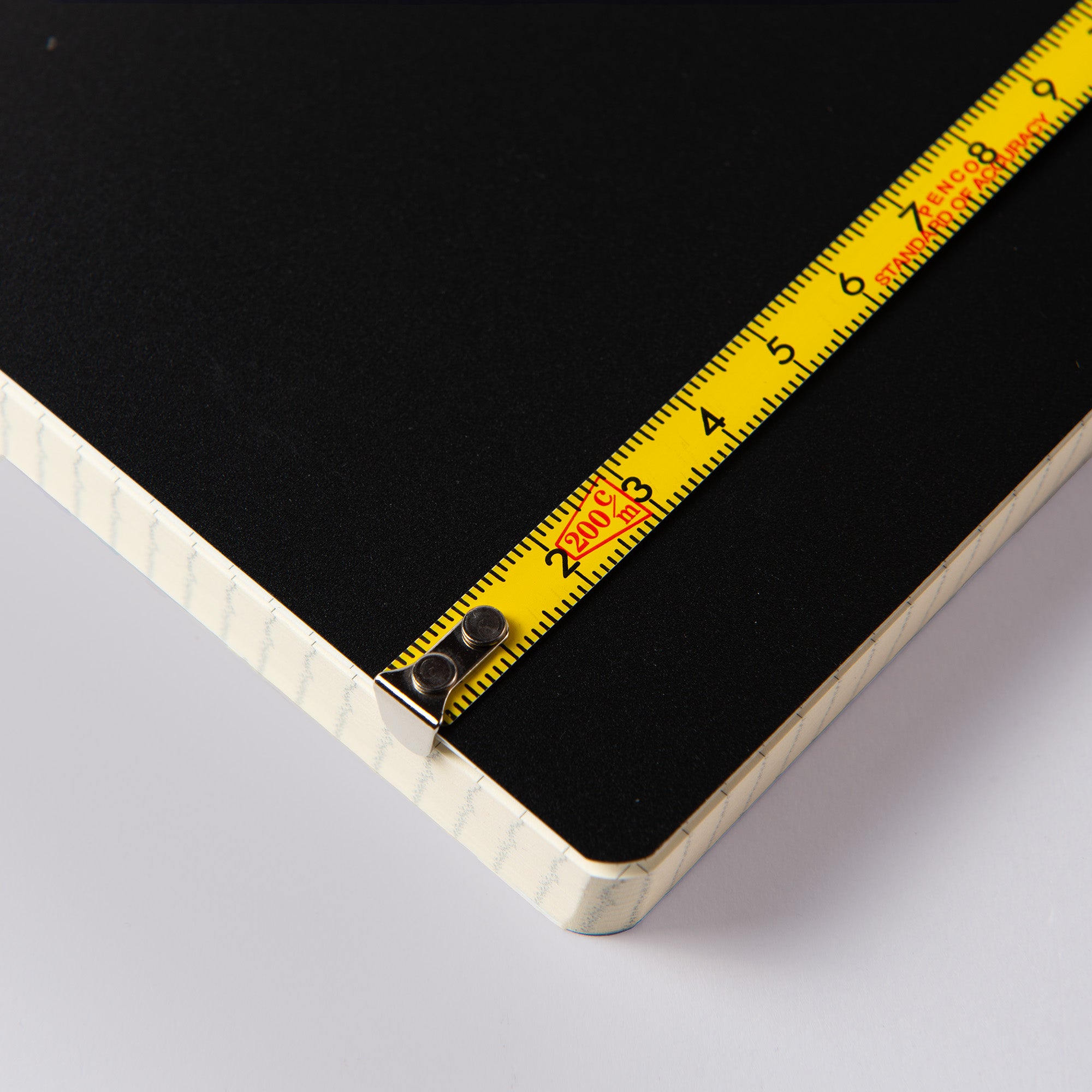Ein gelbes Penco Pocket-Maßband liegt diagonal auf einer schwarzen Oberfläche und zeigt Zahlen und Markierungen zur Genauigkeit. Die Oberfläche ist Teil eines größeren Gegenstands mit einem weißen, strukturierten Rand, was darauf hindeutet, dass es sich um ein Buch oder ein Brett handeln könnte.