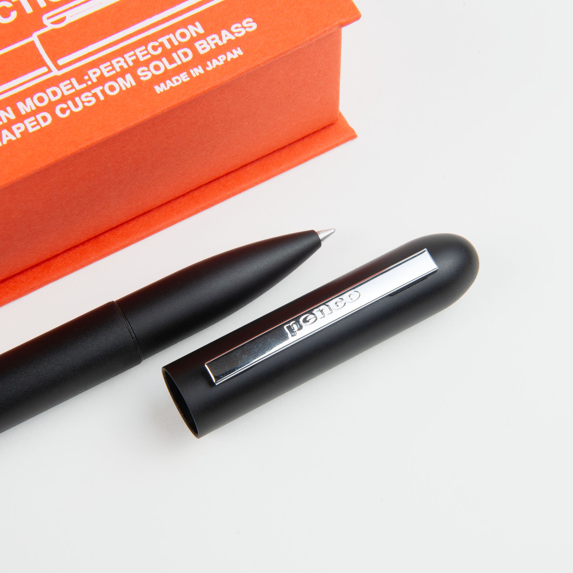 Ein Kompakt-Kugelschreiber - Bullet Pen schwarz von paperlux.store liegt mit abgenommener Kappe neben einer orangefarbenen Schachtel mit weißem Text auf einer weißen Fläche. Hergestellt in Japan.