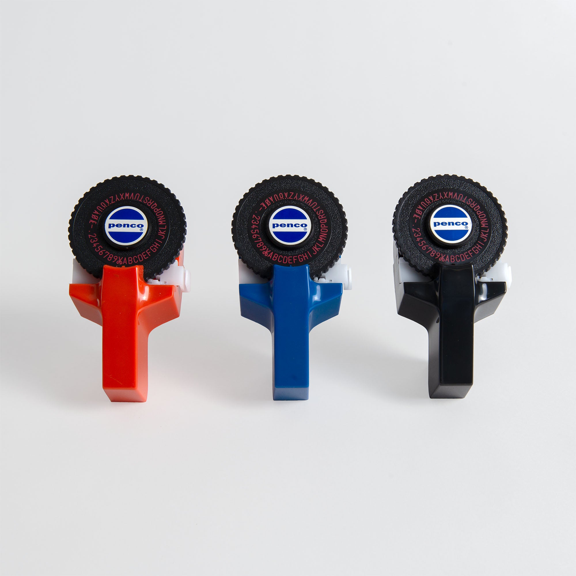 Drei Penco Tape Writer Handauszeichner in den Farben Rot, Blau und Schwarz sind nebeneinander auf einer weißen Fläche angeordnet. Jedes Gerät verfügt über ein drehbares Buchstabenrad, in dessen Mitte der Markenname Penco sichtbar ist.