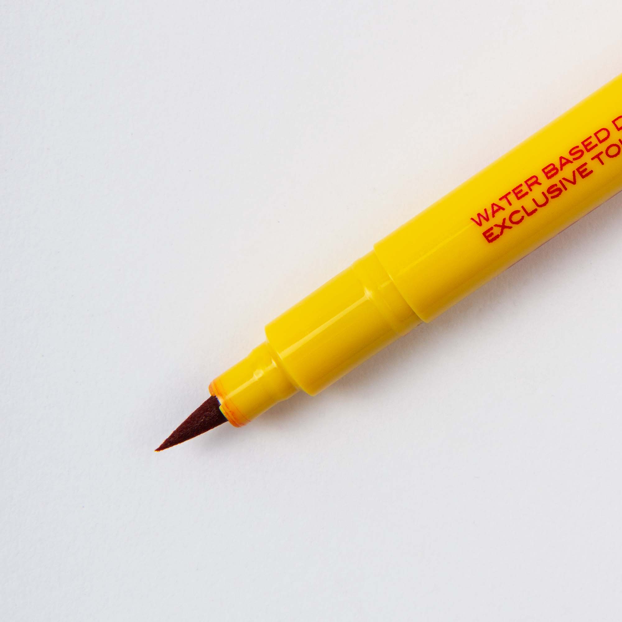 Nahaufnahme eines gelben Brushwriter - Kalligrafiestiftes von Penco mit einer feinen braunen Spitze auf weißer Oberfläche. Auf dem Schaft ist ein roter Text zu sehen, der auf die wasserbasierten und ungiftigen Eigenschaften hinweist und die hochwertige Handwerkskunst hervorhebt.