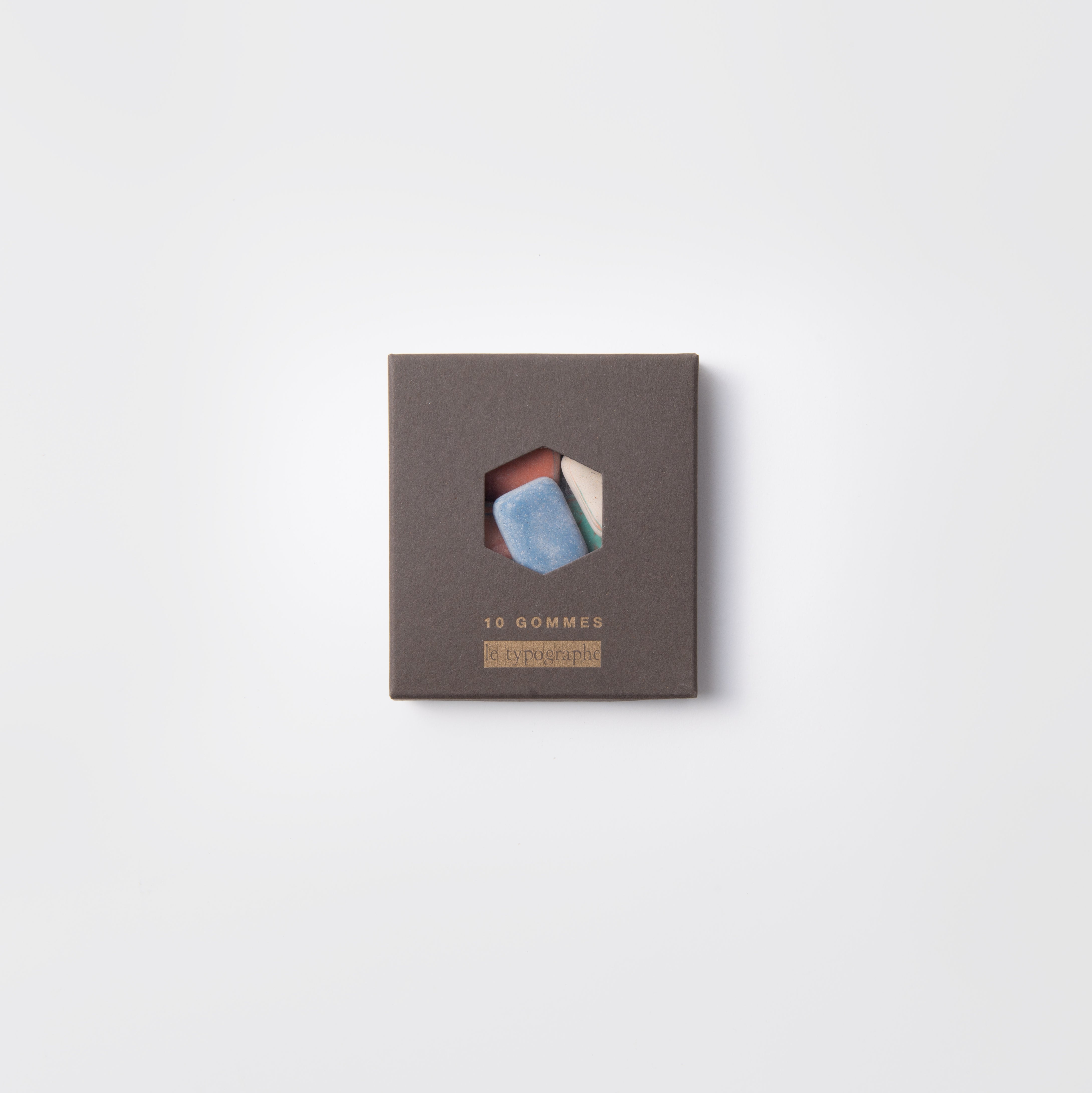 Eine quadratische graue Letterpress-Box mit einem sechseckigen Fenster zeigt bunte Radiergummis "Kieselsteine" von Le Typographe. Auf der Schachtel steht "10 GOMMES" und "le typographe" in Gold, vor einem schlichten weißen Hintergrund.