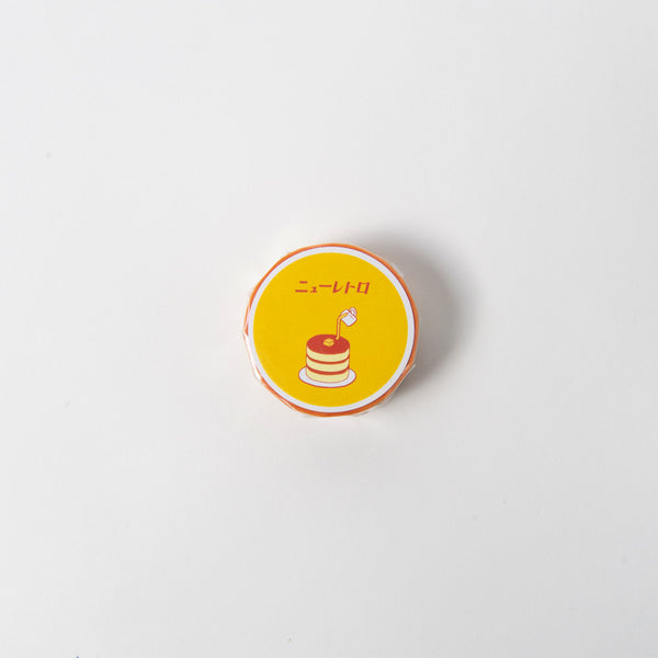 Das Hightide Klebeband - Masking Tape besticht durch sein rundes Design mit gelbem Etikett, das einen Pfannkuchenstapel mit einer Flagge darstellt. Darüber erscheint japanischer Text auf weißem Hintergrund, eingerahmt von einem orangefarbenen Rand.