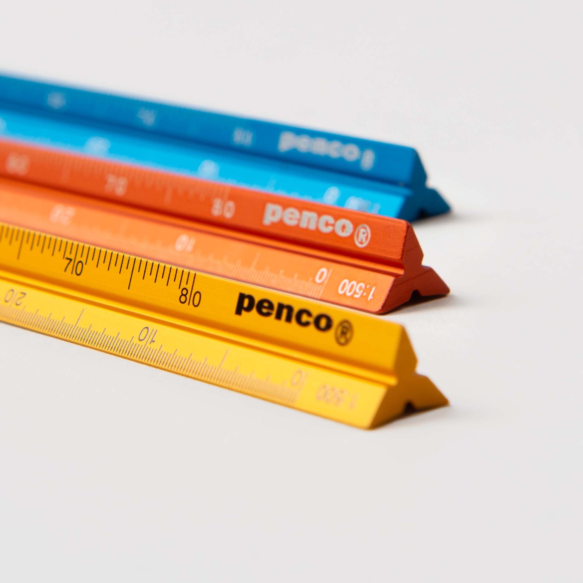 Drei leuchtende Zeichenlineal-Skalenlineal-Lineale von Penco aus robustem Aluminium in Blau, Orange und Gelb sind diagonal auf einer weißen Oberfläche angeordnet. Jedes ist mit „Penco“-Markierungen und Millimeterskalen versehen und verbindet Stil mit Präzision.