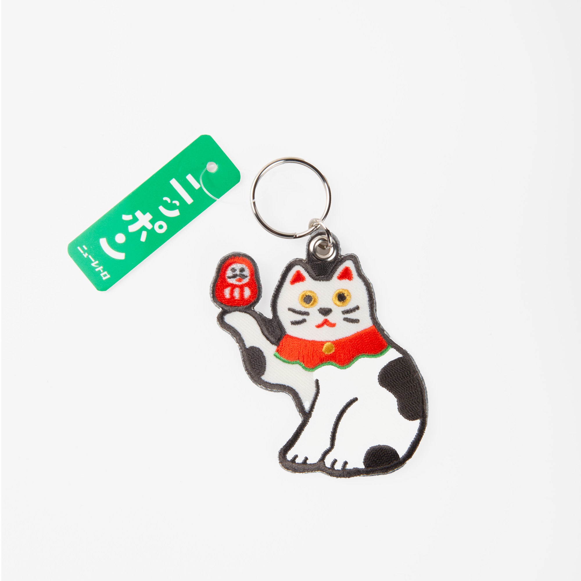 Der Hightide Stitch Work Keychain - Kamera, Daruma & Glückskatze zeigt eine winkende weiße Katze mit schwarzen Punkten und roten Akzenten - eine verspielte Anspielung auf japanische Glücksbringer. Er ist an einem silbernen Ring mit einem grünen Anhänger von Hightide befestigt.