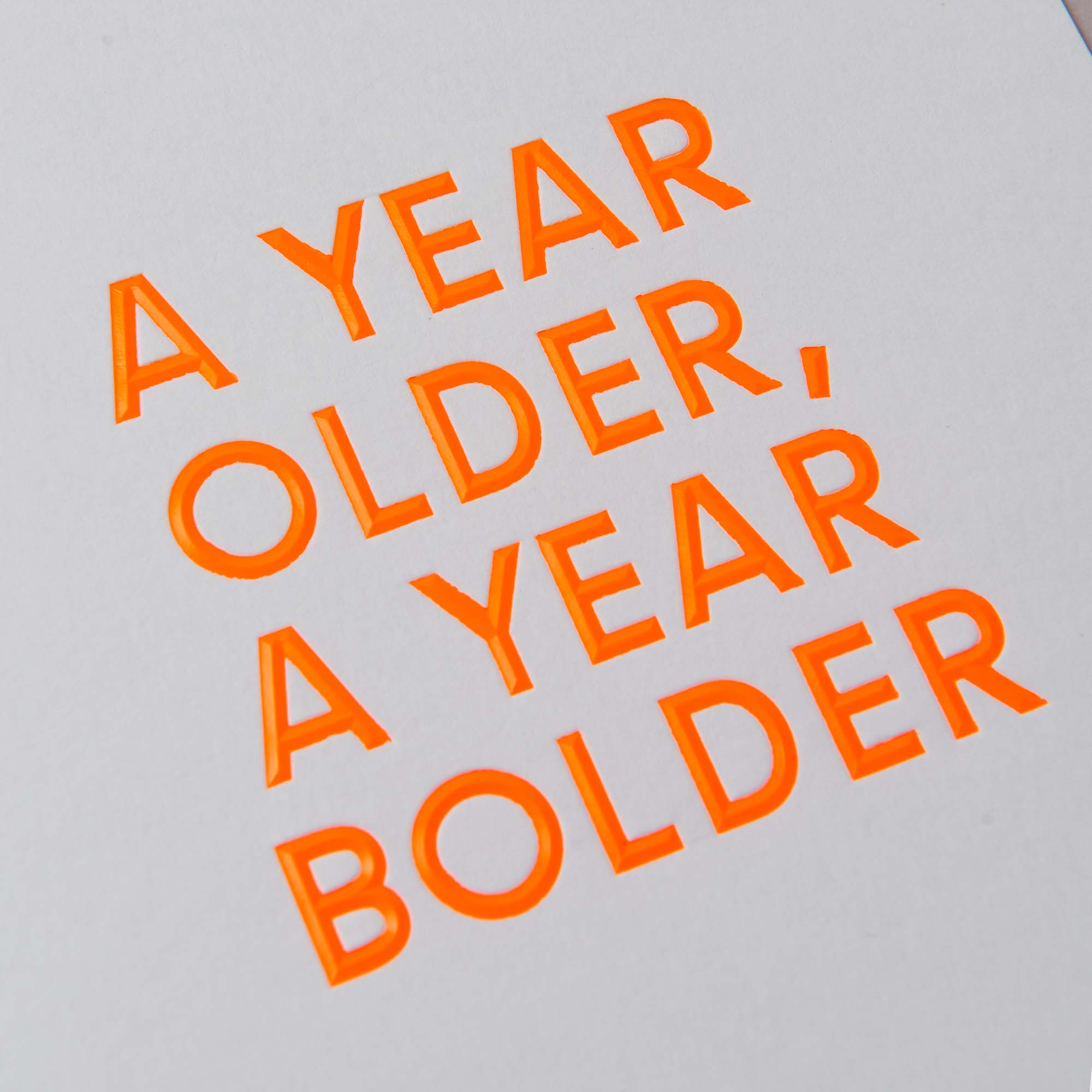 Leuchtend orange Großbuchstaben auf grauem Hintergrund sagen „A YEAR OLDER, A YEAR BOLDER“, ähnlich der Geburtstagskarte „AYOAYB“ von paperlux.store.