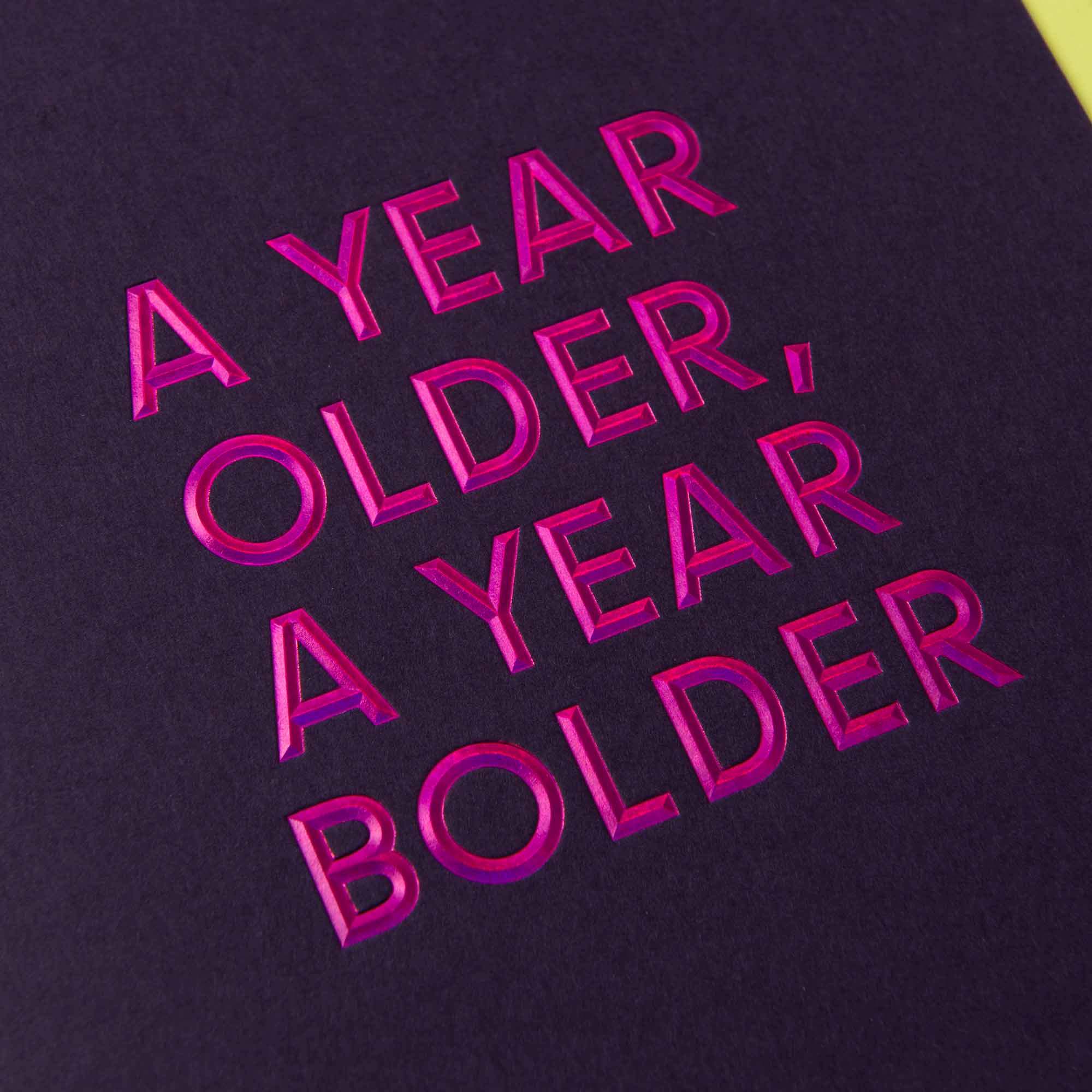 Das Bild zeigt einen dunklen Hintergrund und den kräftigen rosa Text „A YEAR OLDER, A YEAR BOLDER“, der der stilvollen Geburtstagskarte „AYOAYB“ mit Heißfolienprägung von paperlux.store für einen auffälligen Look ähnelt.