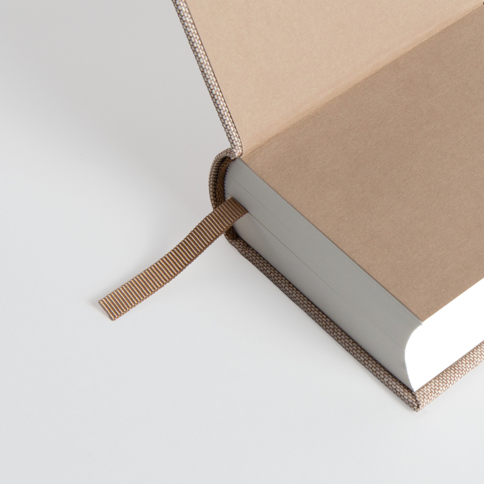 Ein teilweise geöffnetes OWAD Hygge Collection Hardcover-Journal von paperlux.store mit einem schlichten braunen Einband und einem Lesezeichen aus Stoff, abgebildet auf einem weißen Hintergrund. Leere Jahresbegleiter Seiten sind innen sichtbar.