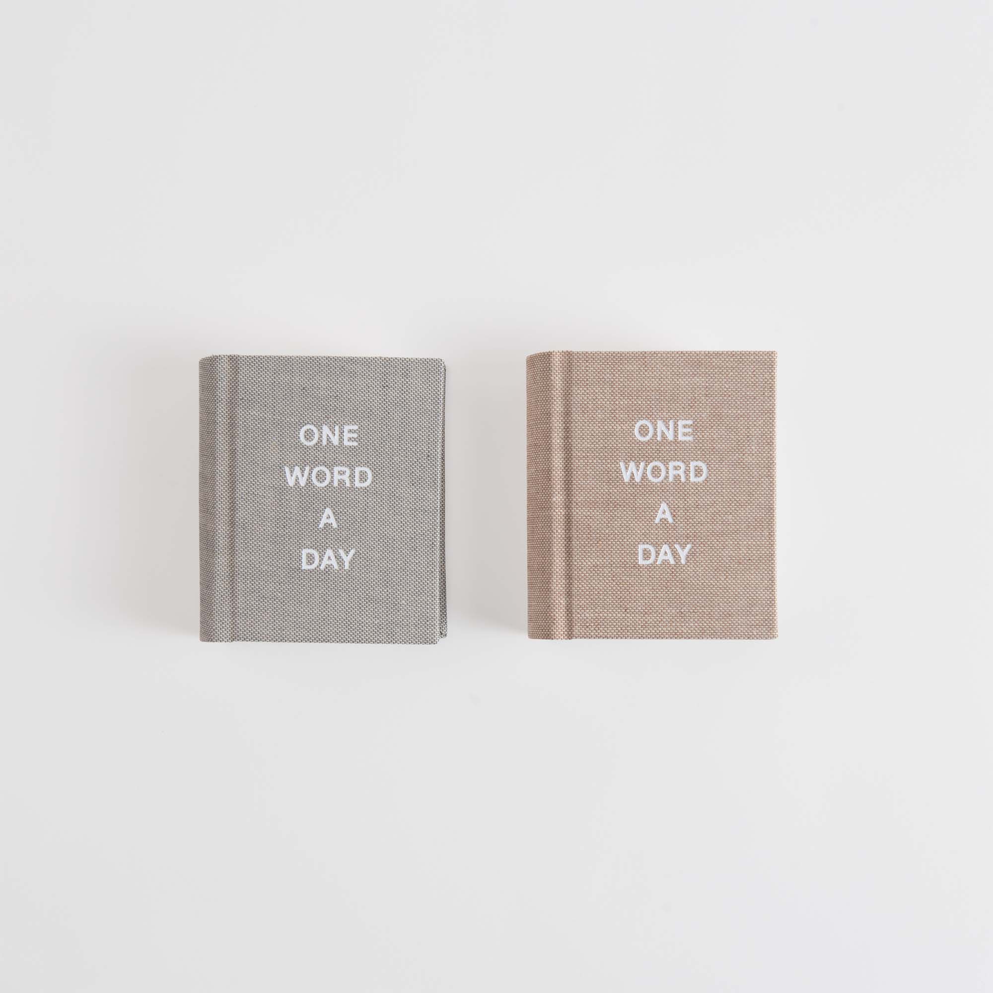Zwei Bücher der OWAD Hygge Collection von paperlux.store stehen nebeneinander auf weißem Hintergrund: eines grau, eines beige. Auf beiden steht "ONE WORD A DAY" in weißen Großbuchstaben, was sie zu perfekten Jahresbegleitern macht.