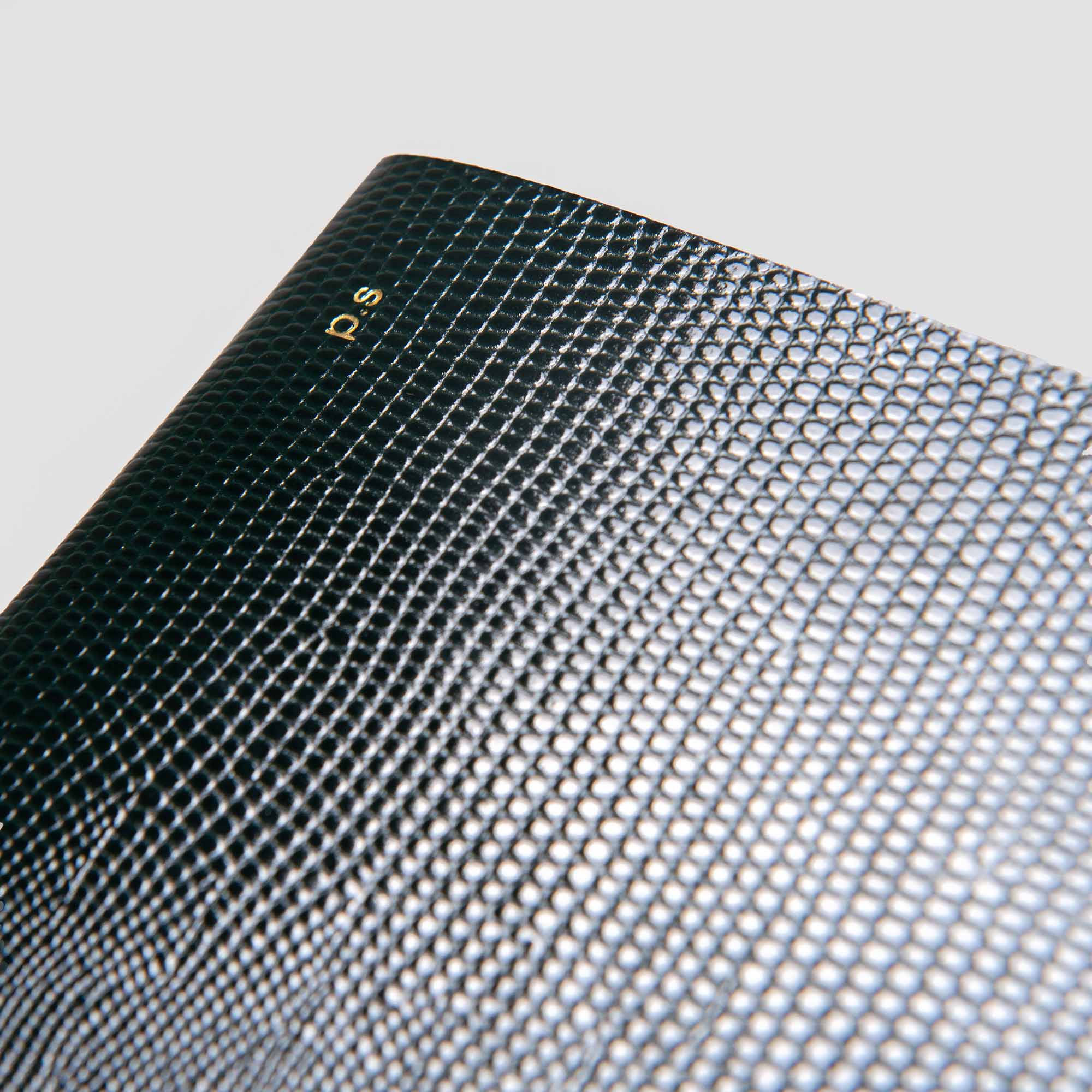 Nahaufnahme von B96 Moss Green Softcover von paperlux.store, mit einer strukturierten Lederoptik-Oberfläche mit kleinen erhabenen Mustern. Die Initialen p.s sind in Gold in der oberen linken Ecke geprägt. Der Hintergrund ist einfarbig hellgrau.