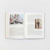 Das aufgeschlagene Buch „Der Savile Row Anzug“ vom Gestalten Verlag zeigt auf der linken Seite eine Straßenszene mit Gebäuden und einer roten Flagge, möglicherweise in der Nähe der Savile Row. Auf der rechten Seite ist ein Vintage-Foto eines Mannes im Anzug zu sehen, das klassische Schneidertechniken demonstriert. Beide Seiten enthalten Absatztext.