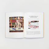 Ein aufgeschlagenes Buch enthüllt den „Savile Row Suit“ des Gestalten Verlags – ein Mann im Anzug sitzt an einem Schreibtisch, dessen Stoffe Handwerkskunst verkörpern. Gegenüber sind Text und gemusterte Stofffotos zu sehen, während im Hintergrund Bücherregale voller Schneidermethoden-Bände stehen.